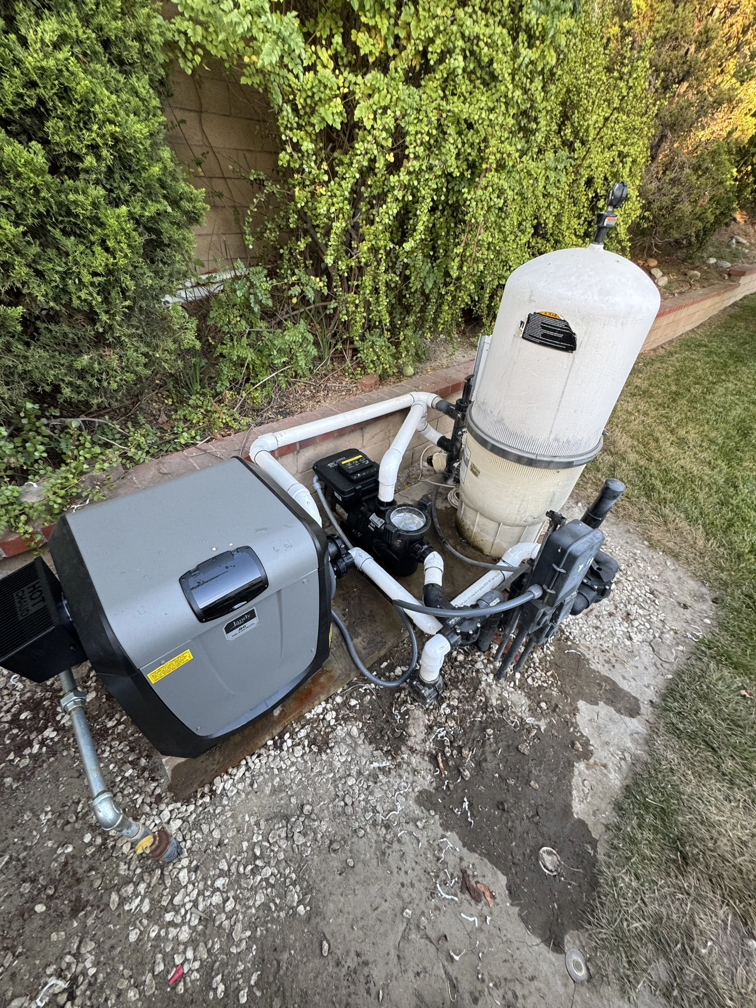 A swimming pool pump system installed outdoors, next to a brick wall and green bushes, with piping and a motorized filter unit.