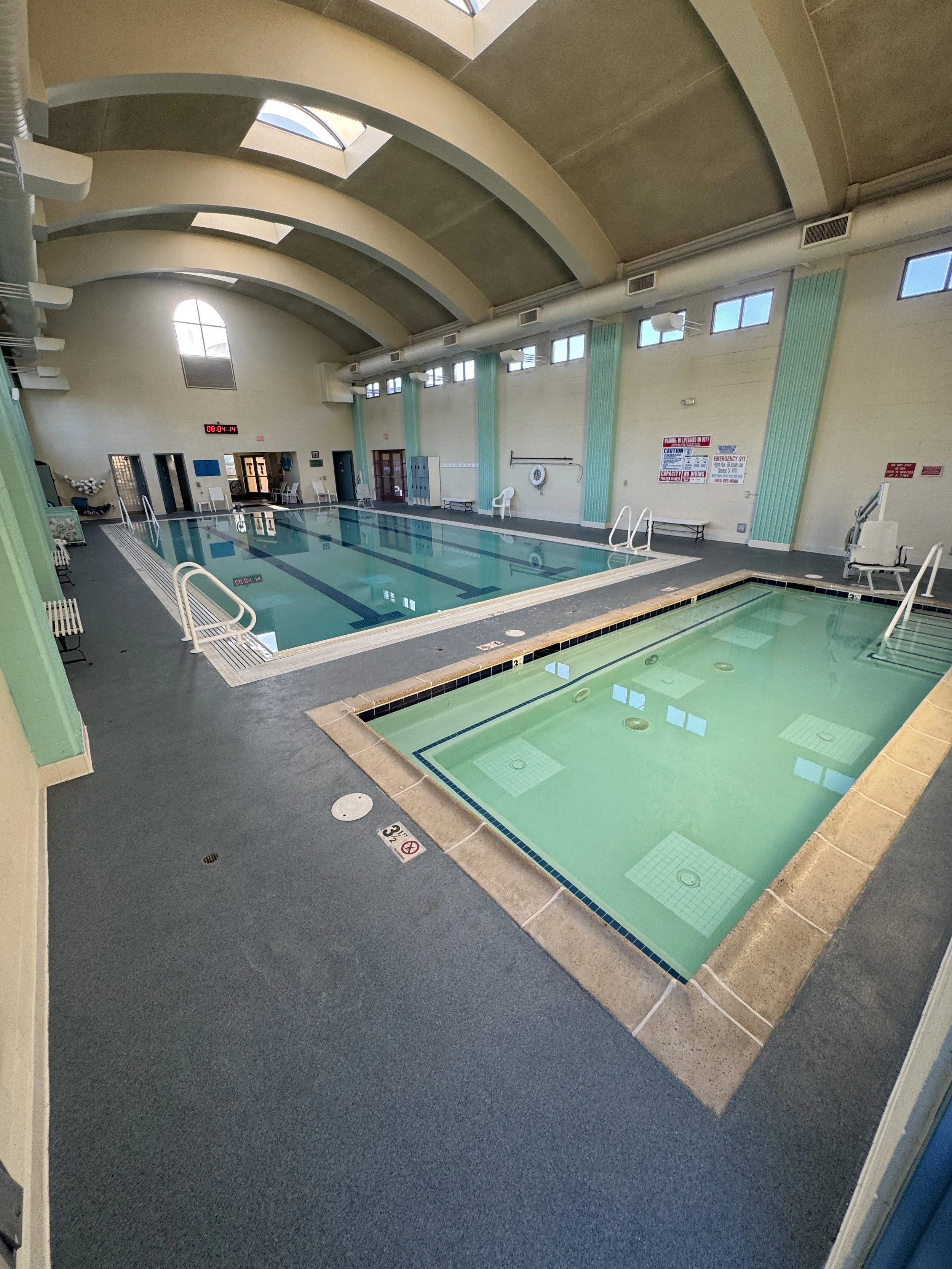 Indoor swimming pool facility with a large main pool and a smaller hot tub, high arched ceiling with windows, and lockers in the background.