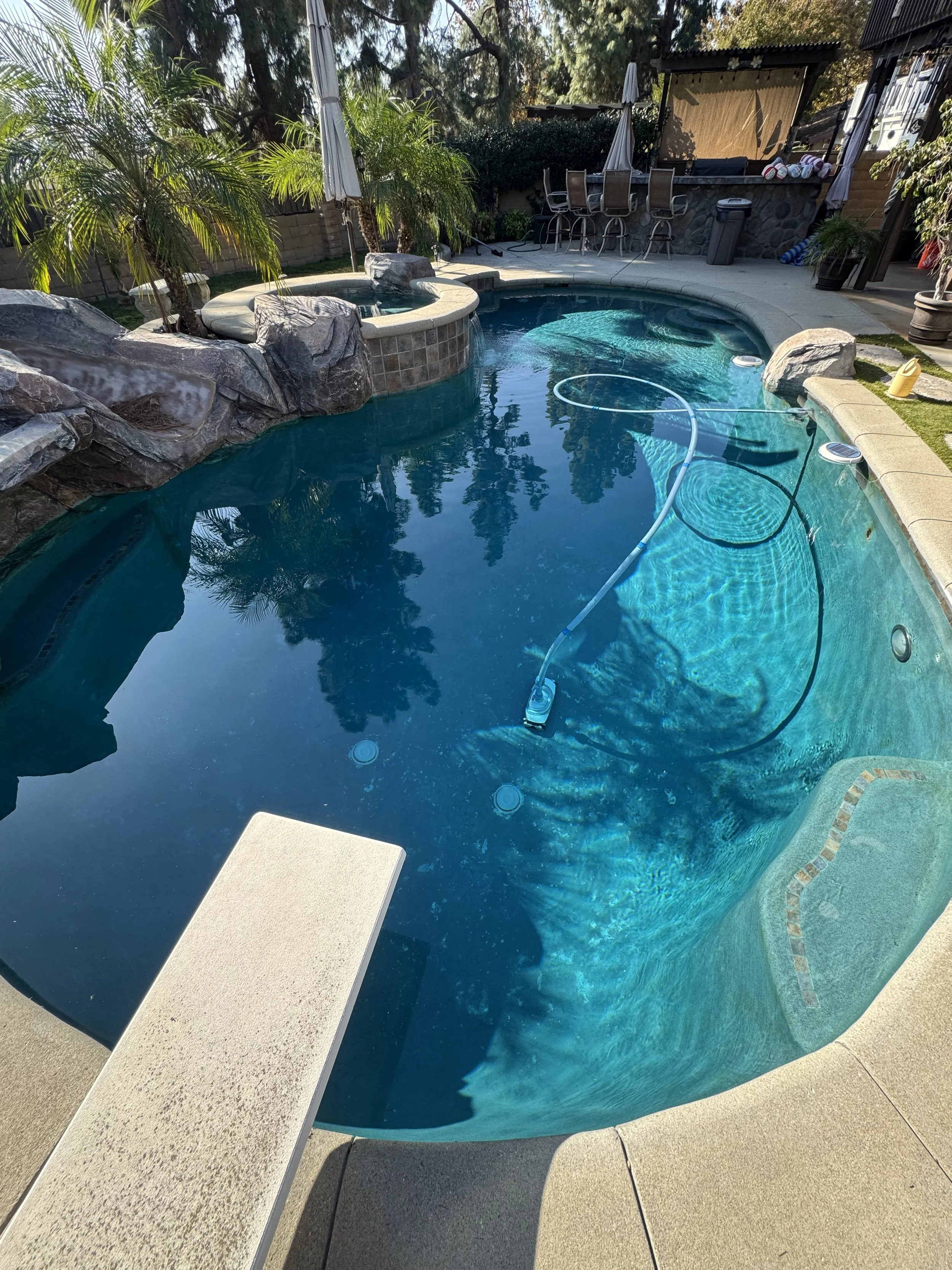 A backyard swimming pool with a small hot tub, surrounded by rocks and palm trees. There is a floating pool cleaning device and a diving board on the pool's edge. In the background, there is outdoor seating with umbrellas and a bar area.