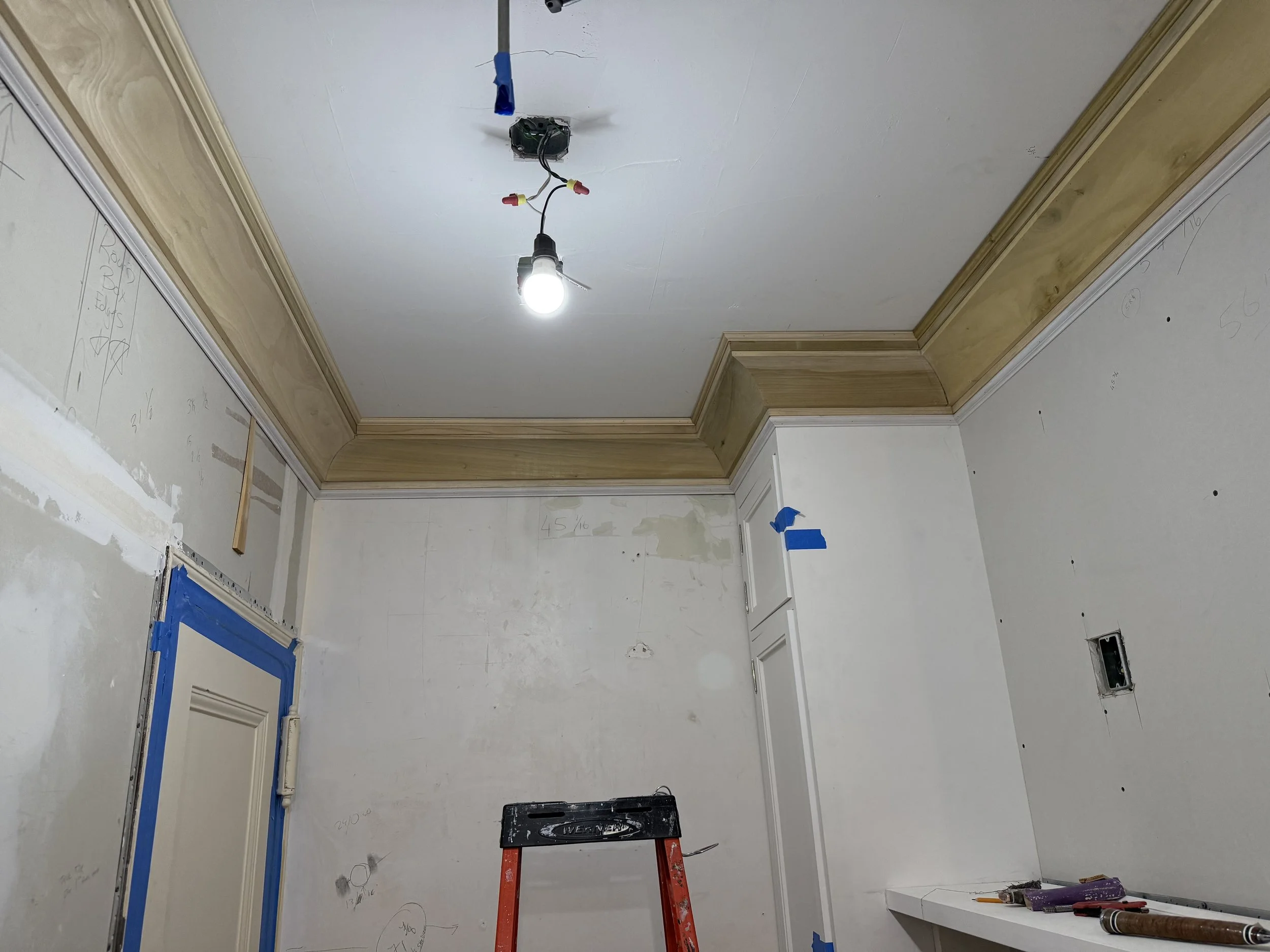 A room under renovation with a white ceiling, wooden crown molding, and construction tools. A hanging light bulb is installed, and blue painter's tape covers the door frame and various areas. An orange ladder is positioned in the center. The walls ha