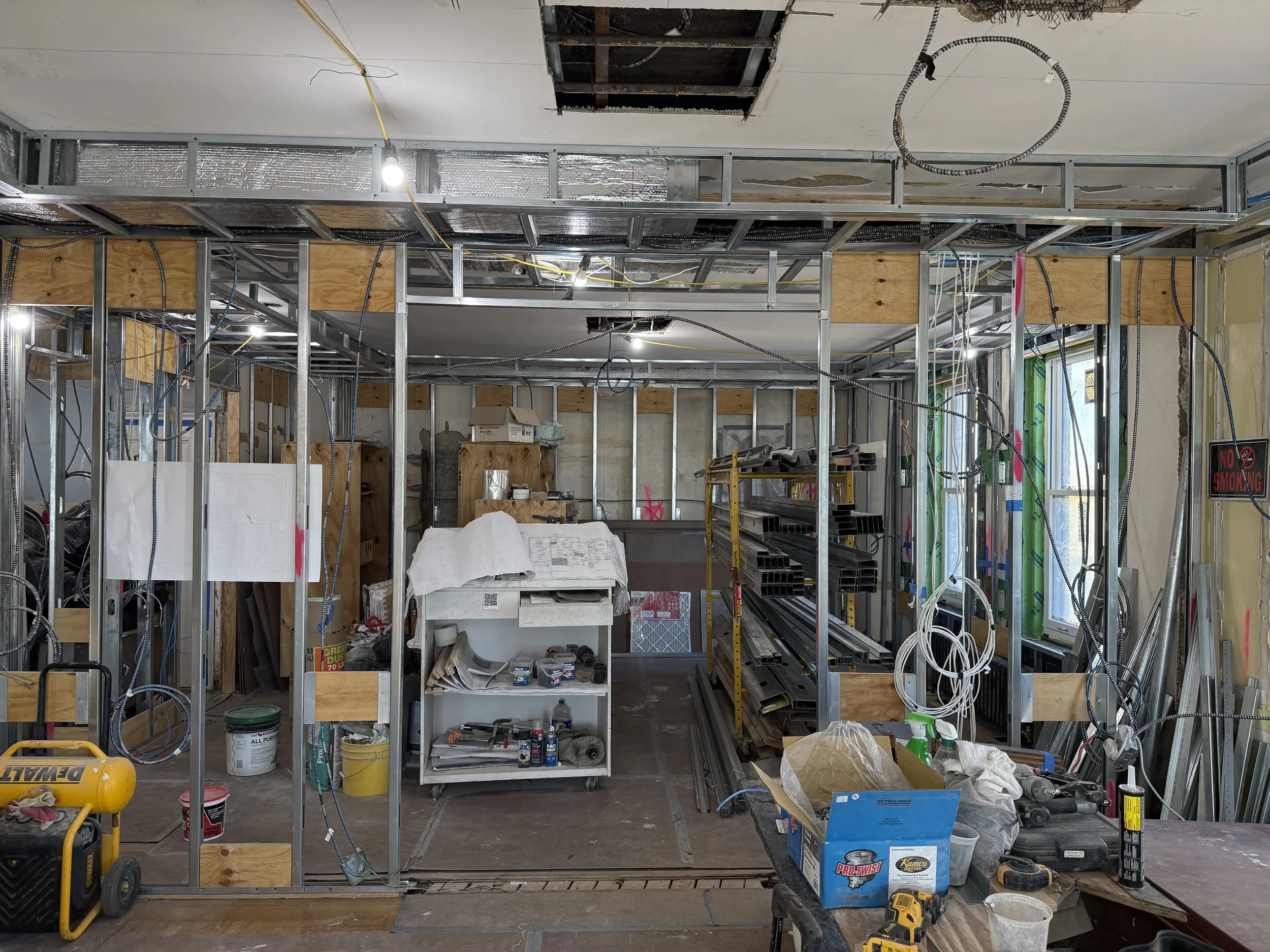 Interior of a building under construction with exposed metal framing, electrical wiring, tools, and building materials scattered around.