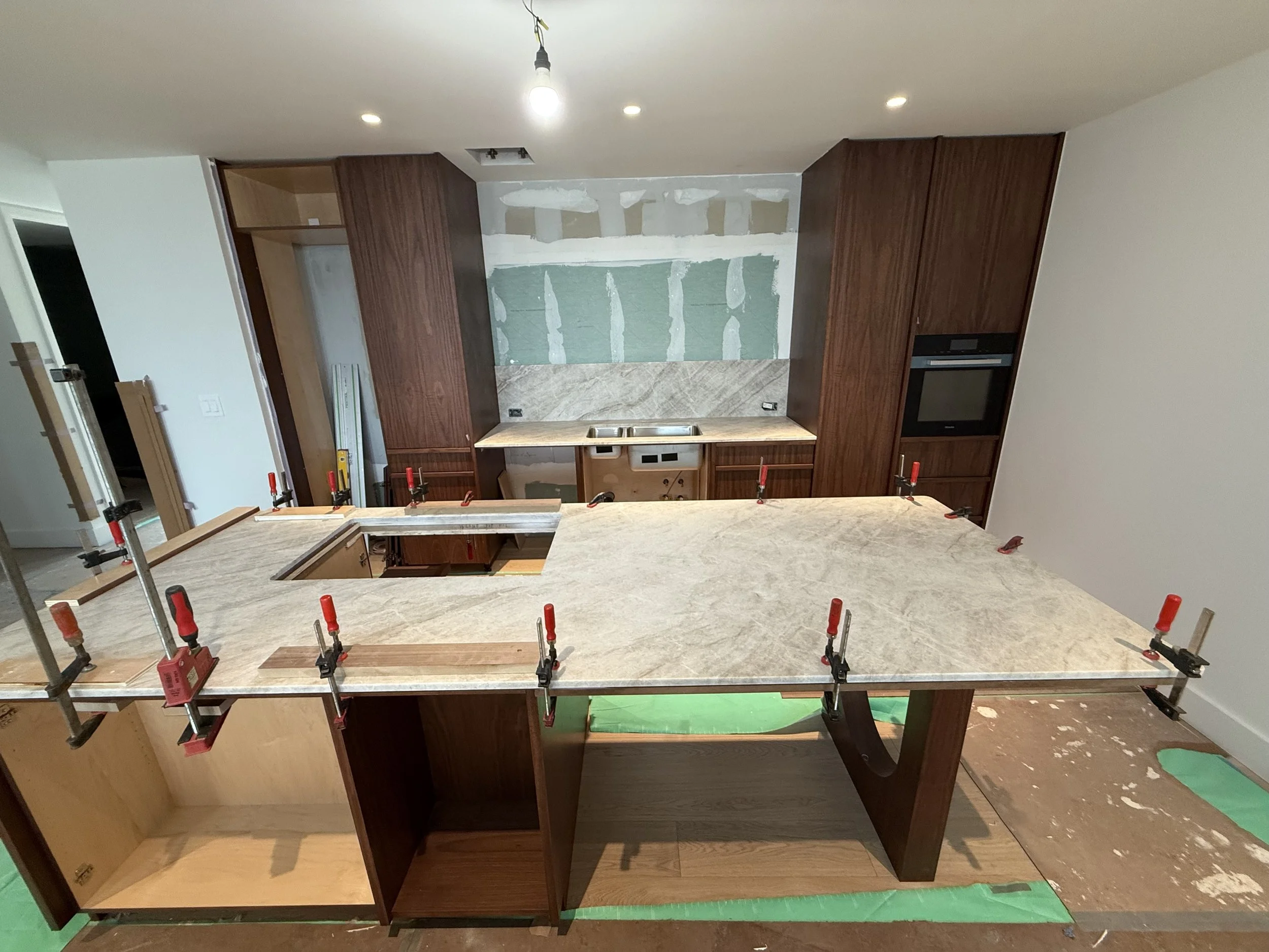 Kitchen under construction with a large island countertop being installed, held in place by clamps. In the background, there are wooden cabinets, a sink, and partial wall drywall.