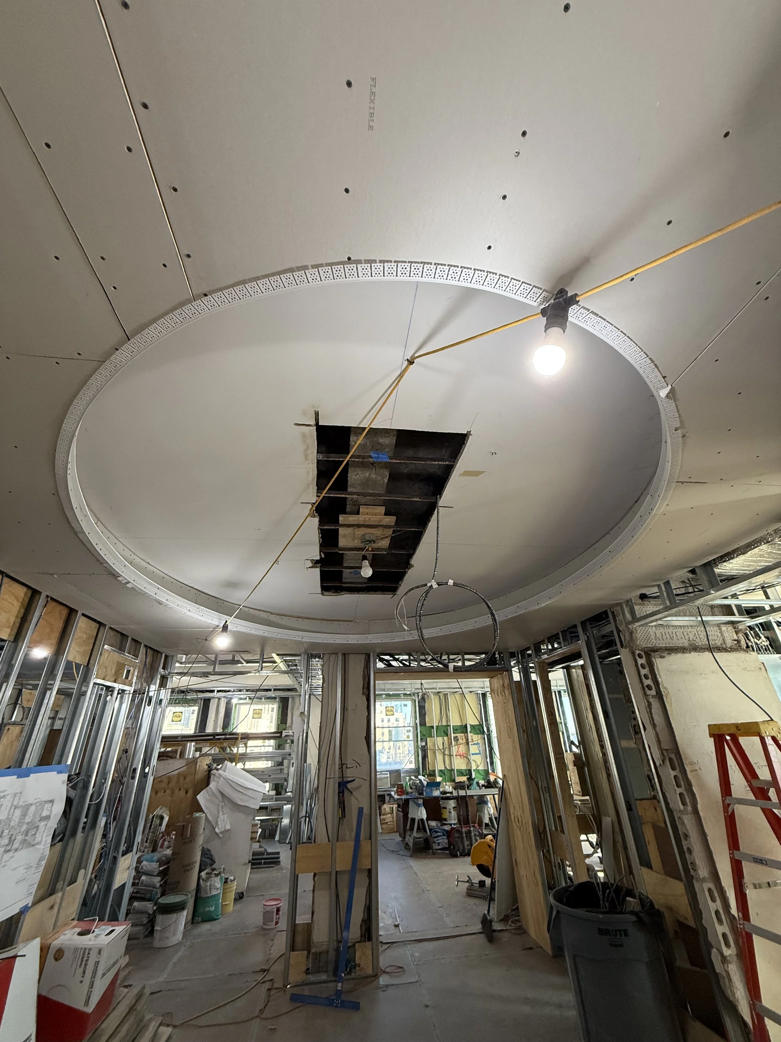 Construction site with drywall ceiling, circular cutout, and hanging lights; exposed framing, tools, and supplies in the background.