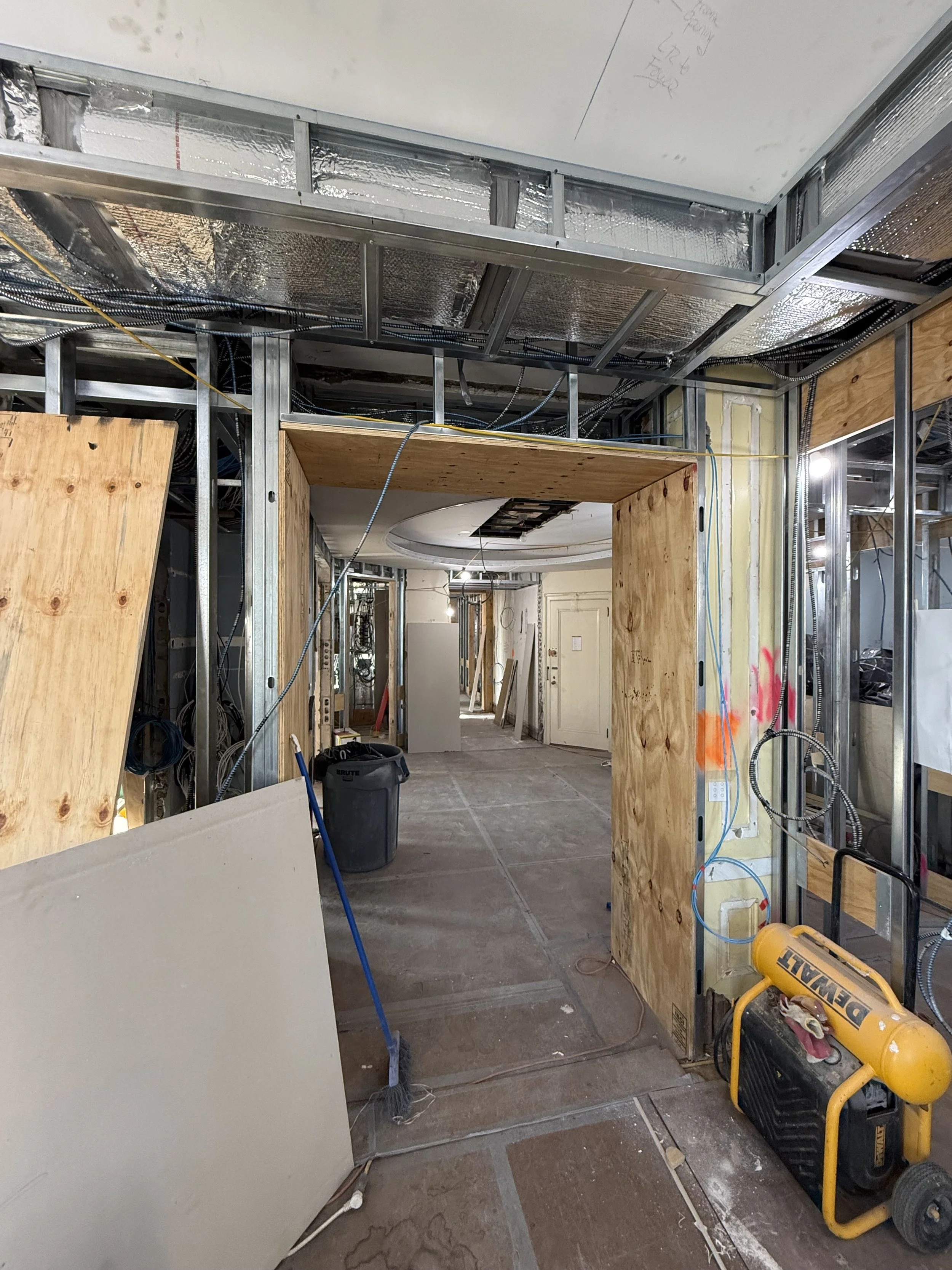 Interior of a building under construction with exposed framing, drywall, and electrical wiring. Plywood partitions create doorways, and construction equipment is visible, including a yellow DeWALT air compressor and a trash bin.
