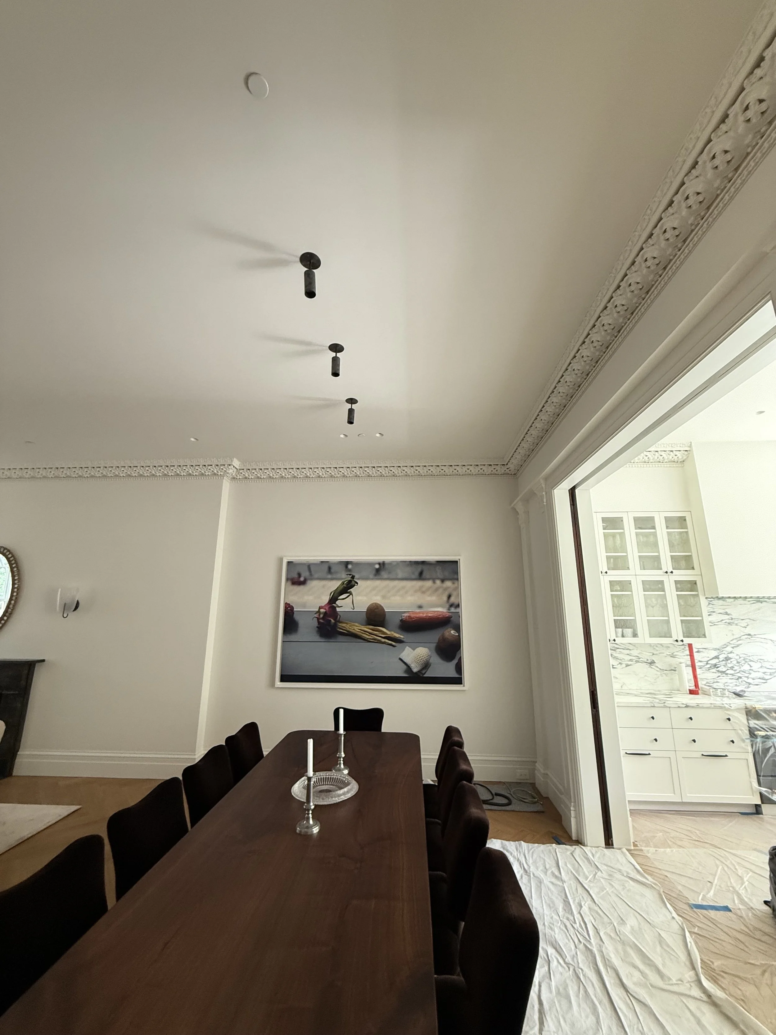 A dining room with a long wooden table, black chairs, and candle holders with white candles. The wall features a framed photo of various objects on a surface. The ceiling has decorative crown molding and three ceiling socket fixtures, with some light