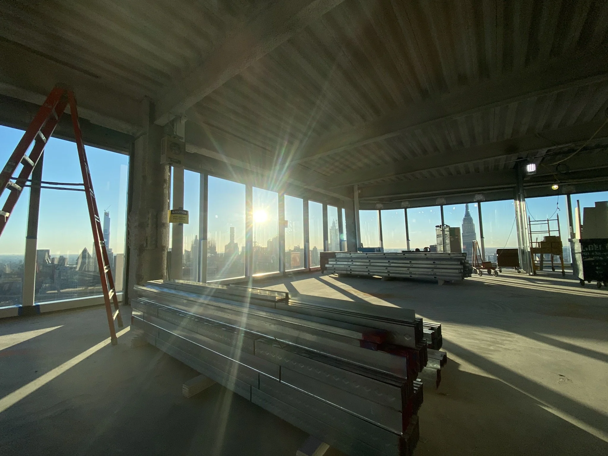 Construction site inside a high-rise building with metal pipes, a ladder, and city skyline view through large windows, with sunlight shining in.