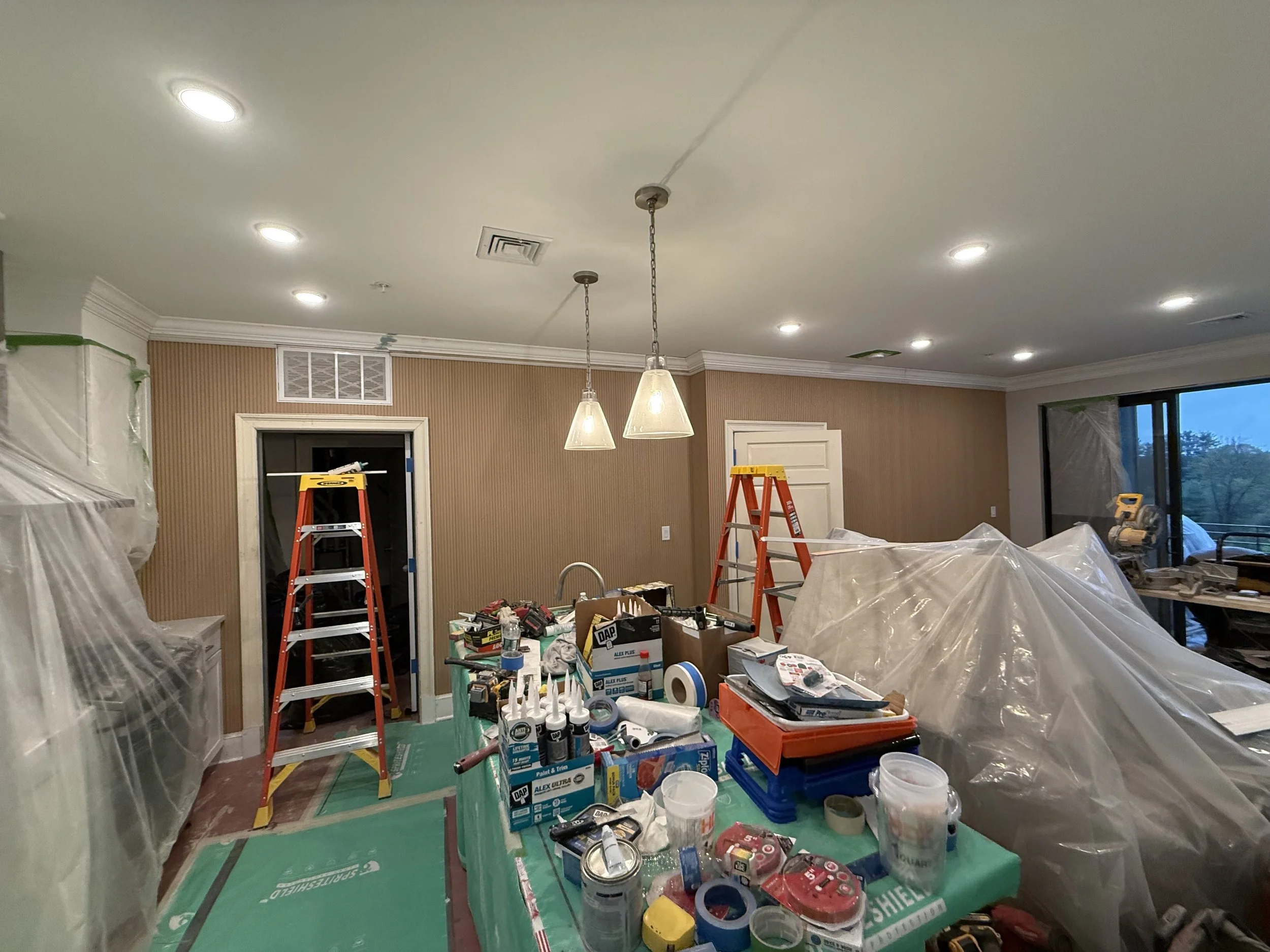 Room under renovation with tools, ladders, plastic coverings, and construction supplies.