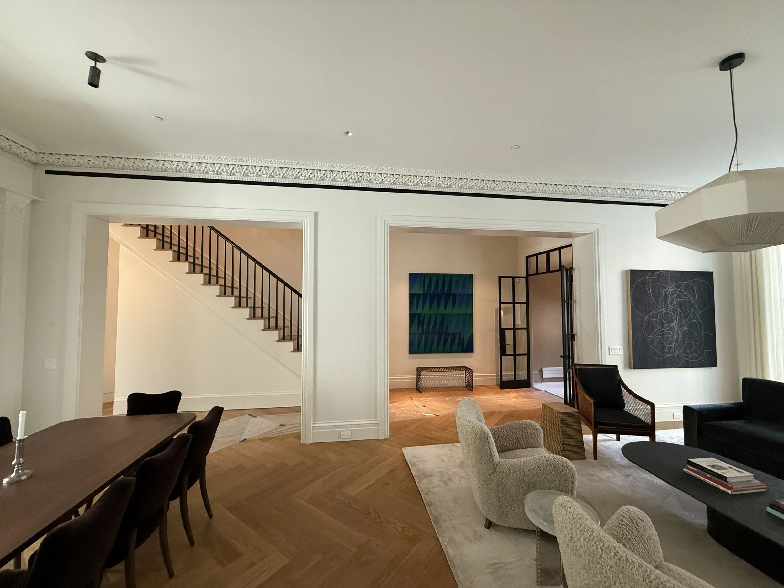Elegant living room with wooden floor, black sofa, beige armchairs, modern artwork, and a staircase leading upstairs.
