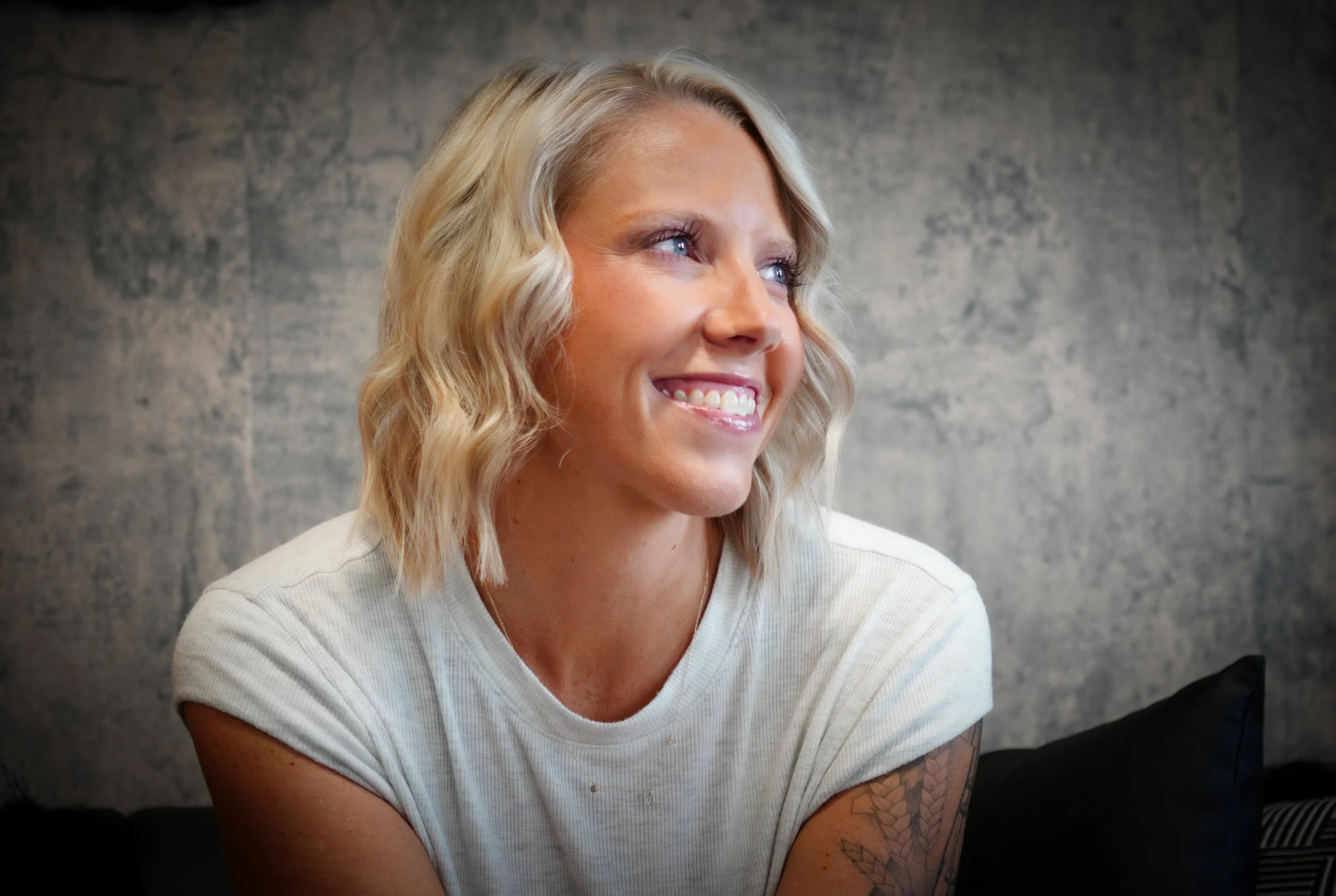 A smiling blonde woman with wavy hair looking to the side, wearing a white t-shirt, seated in front of a gray textured wall.