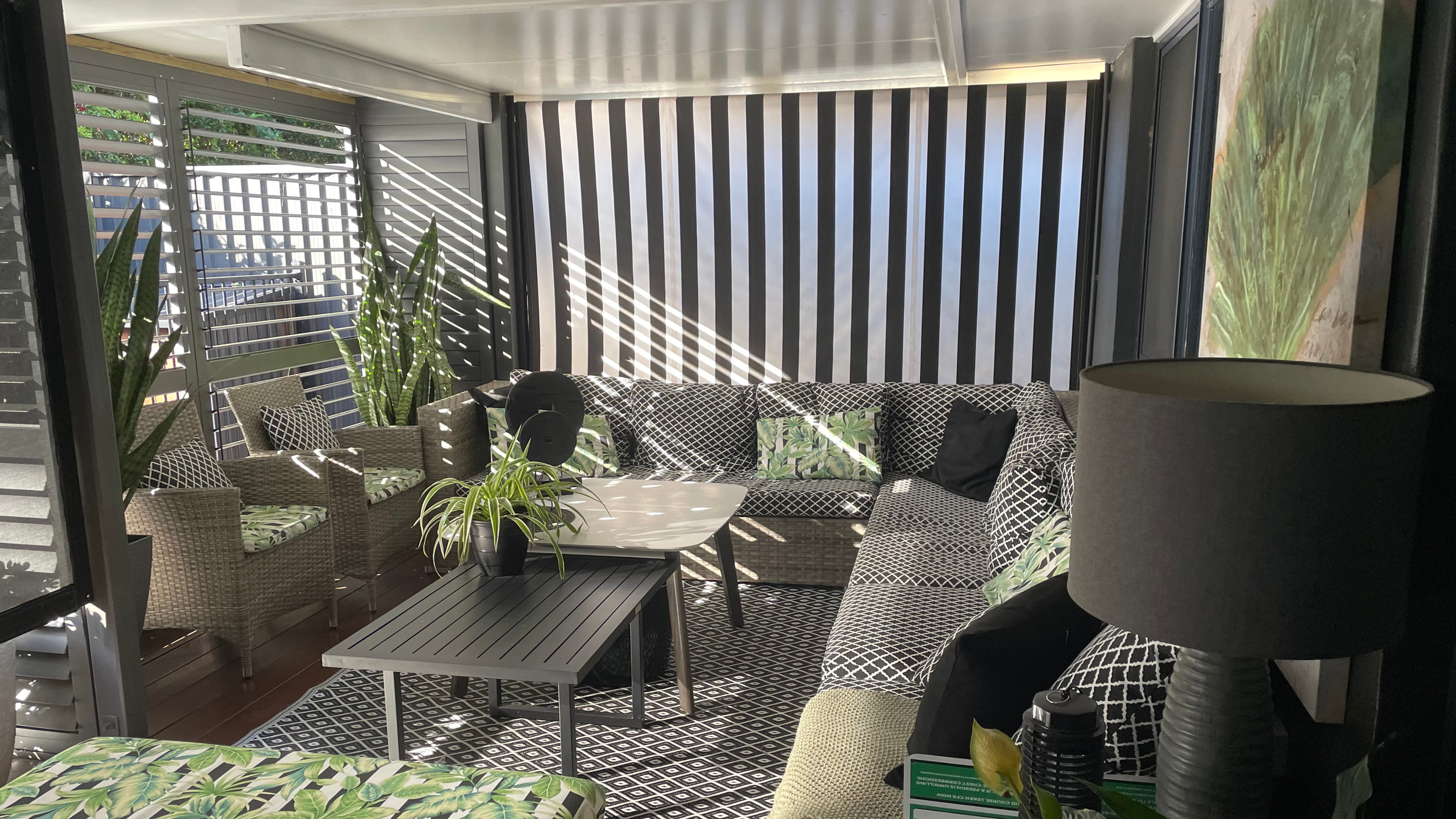 Indoor patio with wicker chairs, a gray sectional sofa with black and green pillows, potted plants, a coffee table, and a patterned rug, with sunlight coming through striped wall panels.