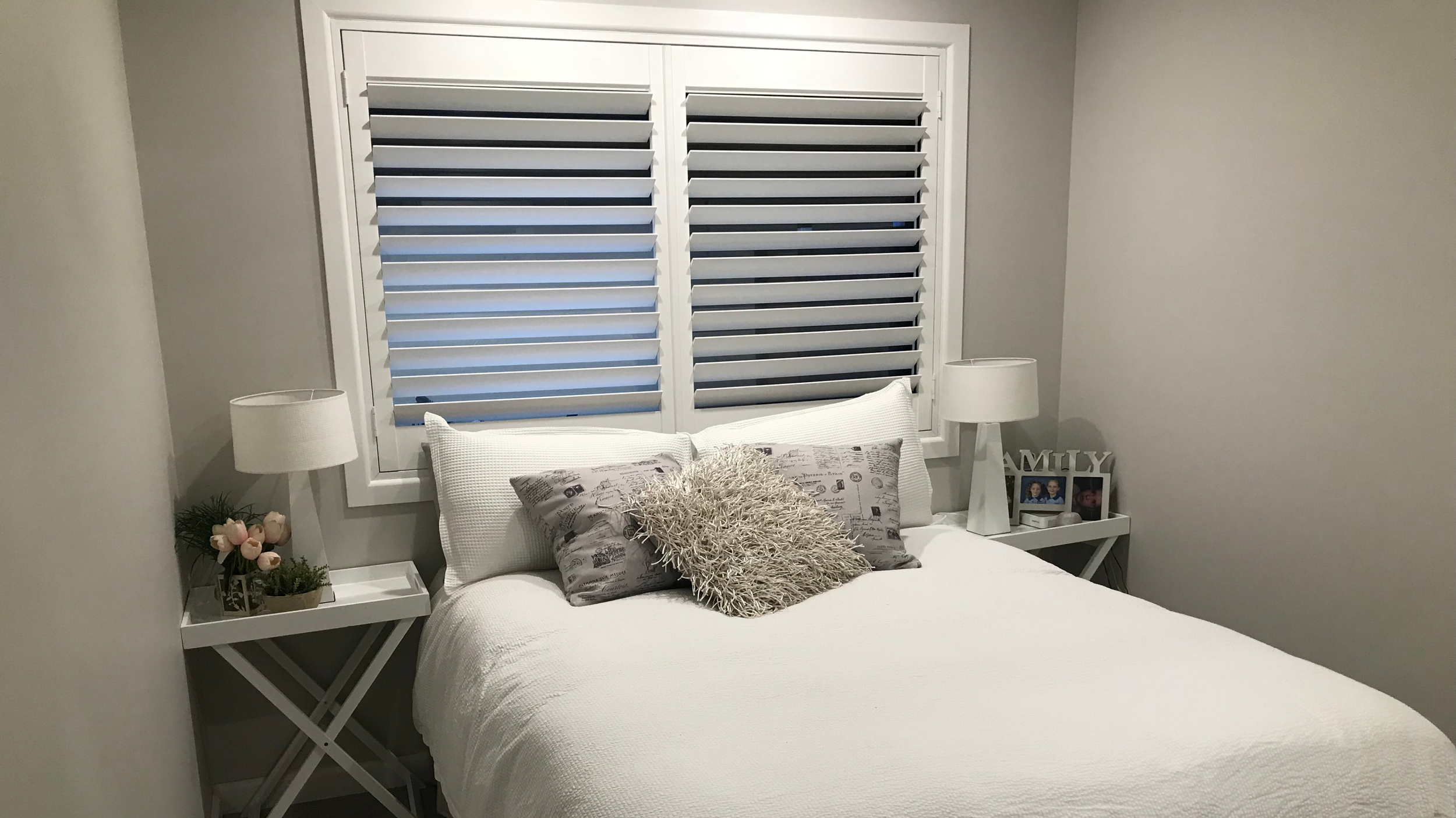 Bedroom with white bed, pillows, and two bedside tables with lamps and decorative items, window with white shutters.