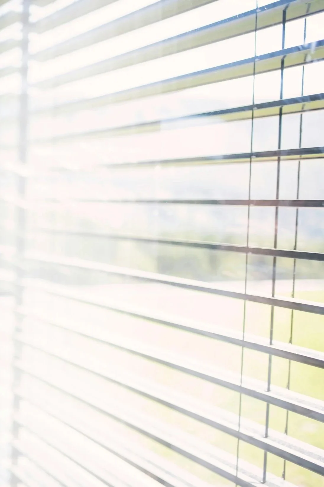 Close-up of window blinds with sunlight shining through, partially obscuring the view outside.