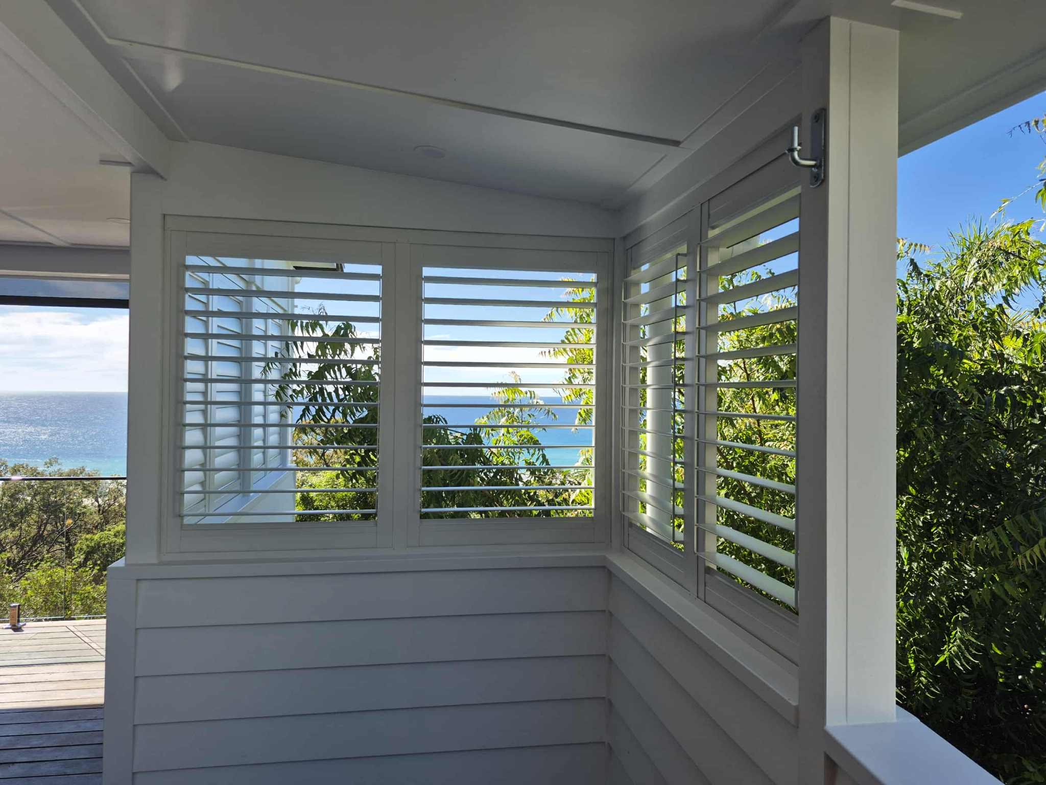 View from inside a bright porch with white plantation shutters, overlooking trees, a wooden deck, and the ocean in the distance.