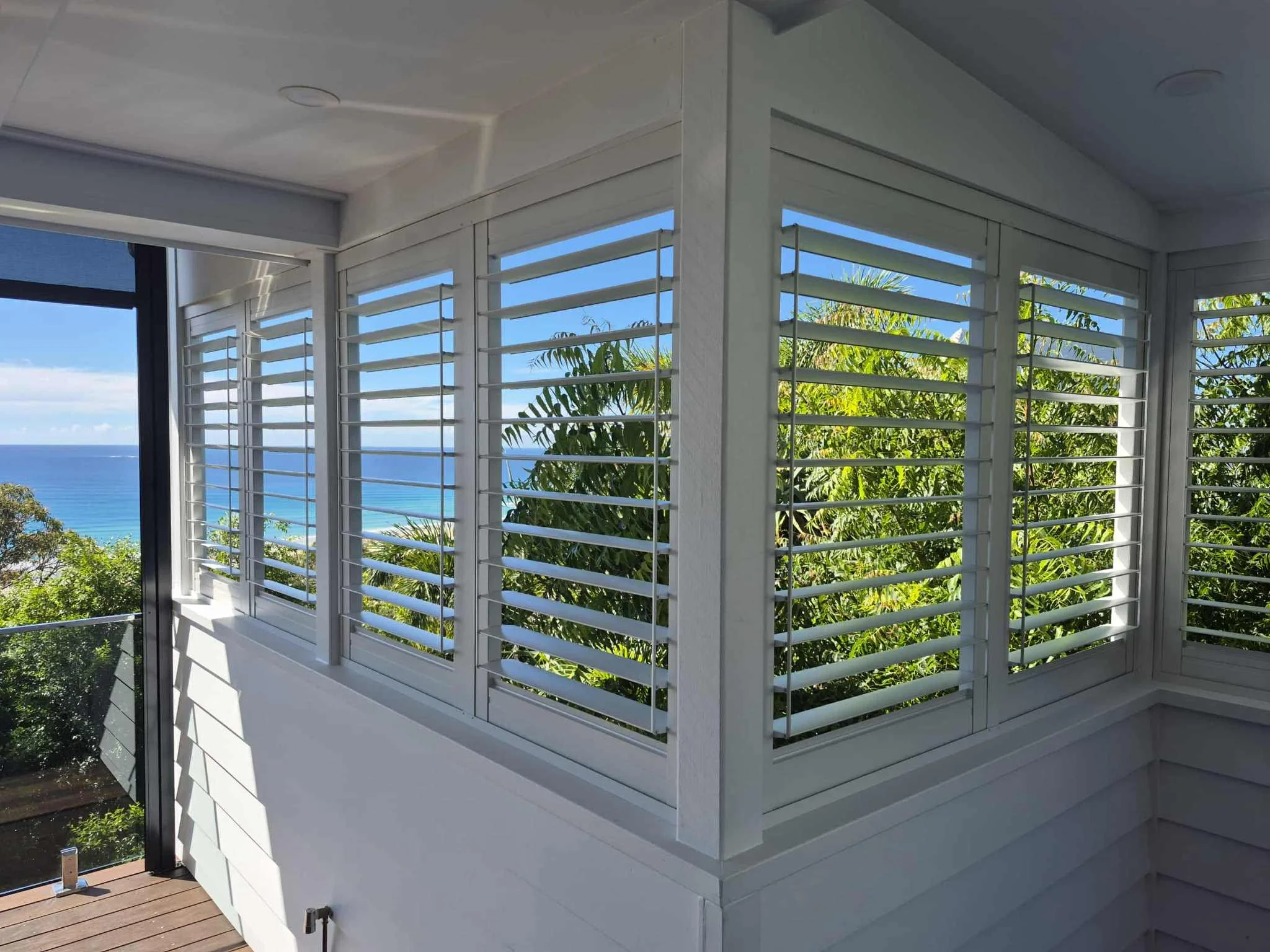 View from a corner of a balcony with white plantation shutters on the windows, overlooking lush green trees and the ocean in the distance on a sunny day.