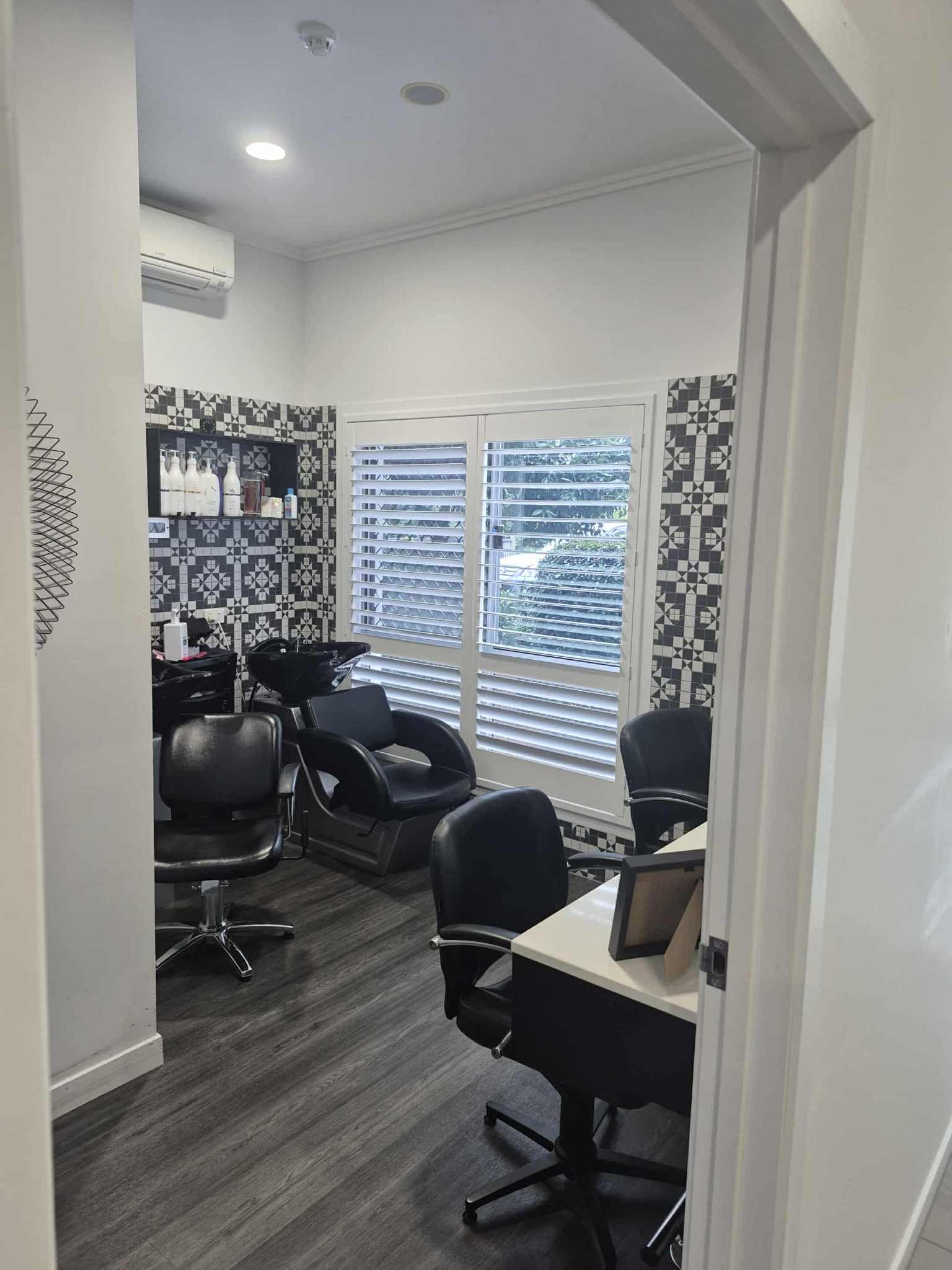 Interior of a hair salon with salon chairs, a shampoo station, a desk with a computer, and large windows with white blinds.