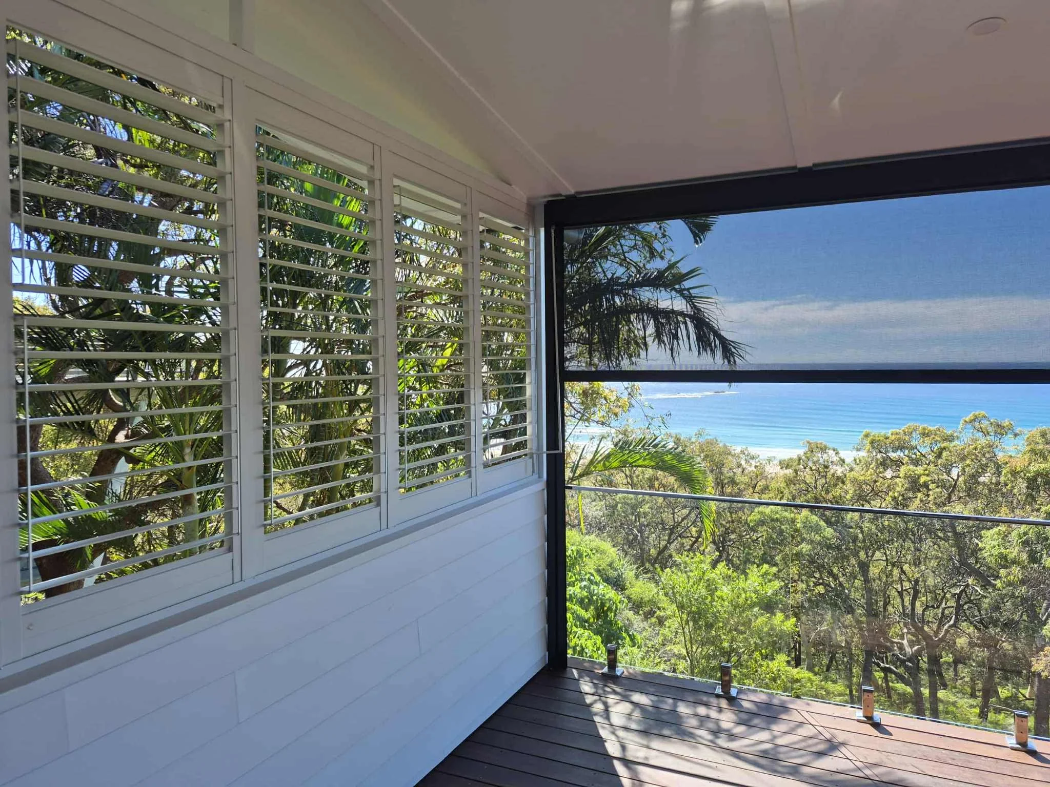 Balcony with glass railing overlooking lush green trees, blue ocean, and clear sky, partially covered by white and black awnings.