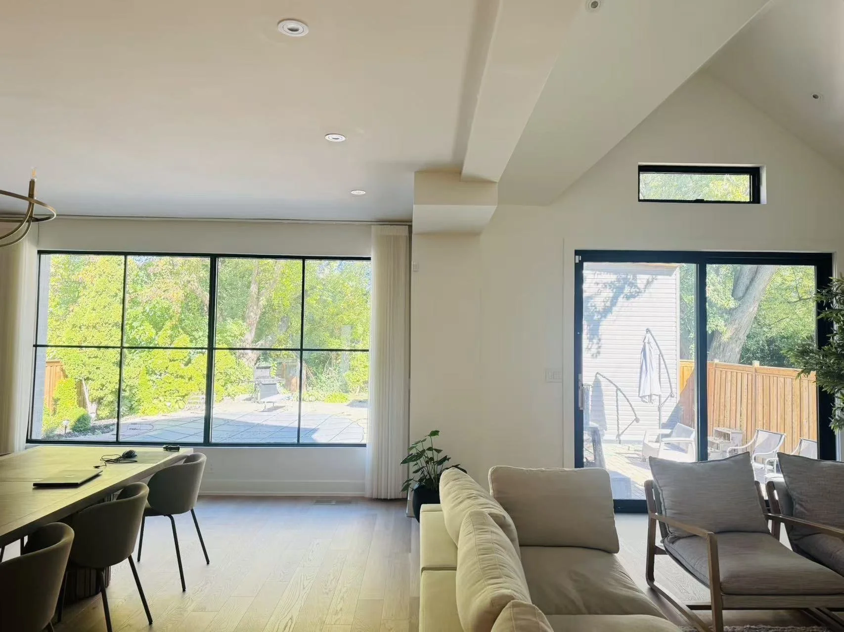 A modern living room with large windows overlooking a backyard with trees. There is a white sofa, wooden chairs, and a dining table with chairs. A sliding glass door leads to an outdoor patio.
