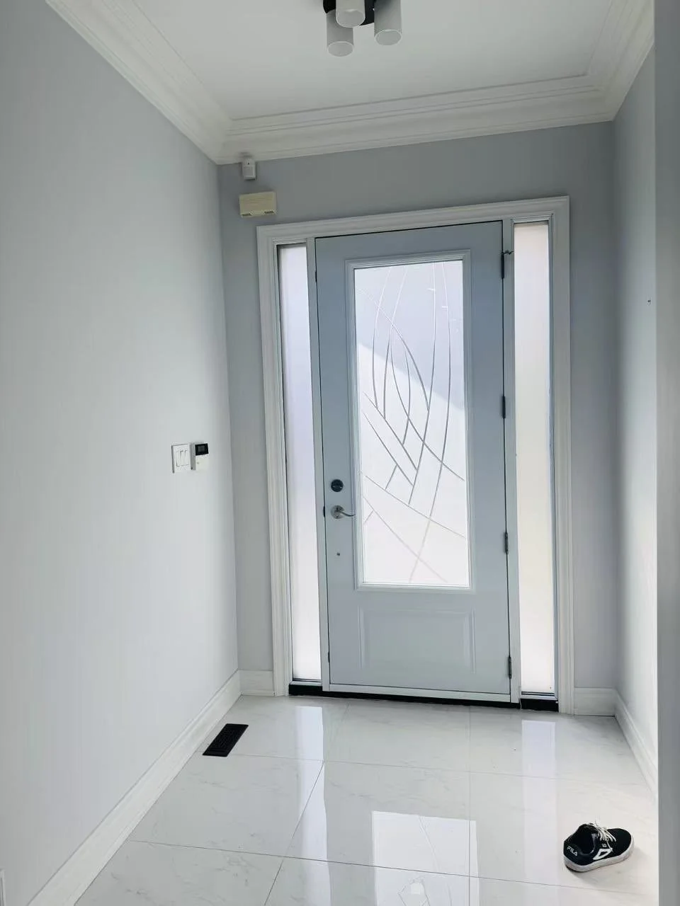 Entrance foyer with a white door featuring decorative frosted glass, a pair of black sneakers on the white tiled floor, and a wall-mounted thermostat next to the door. The room has white walls, crown molding, and a ceiling light fixture.