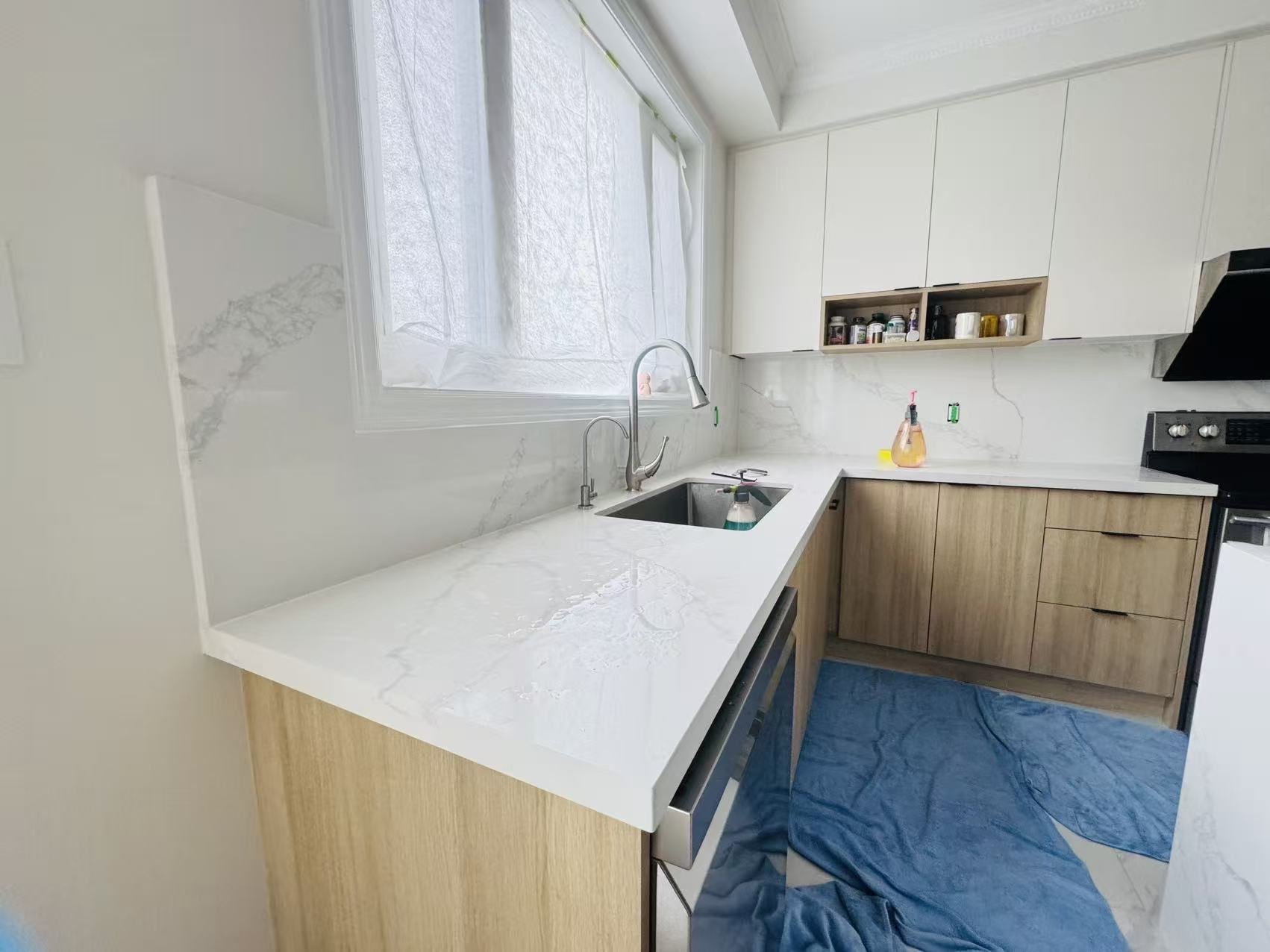 A modern kitchen with white marble countertops, wooden lower cabinets, white upper cabinets, and a large window covered with a plastic sheet. There is a sink with a faucet, a spray bottle, and some cleaning supplies. Shelves with jars and containers are visible above the countertop. A blue towel or cloth is on the floor near the cabinets.