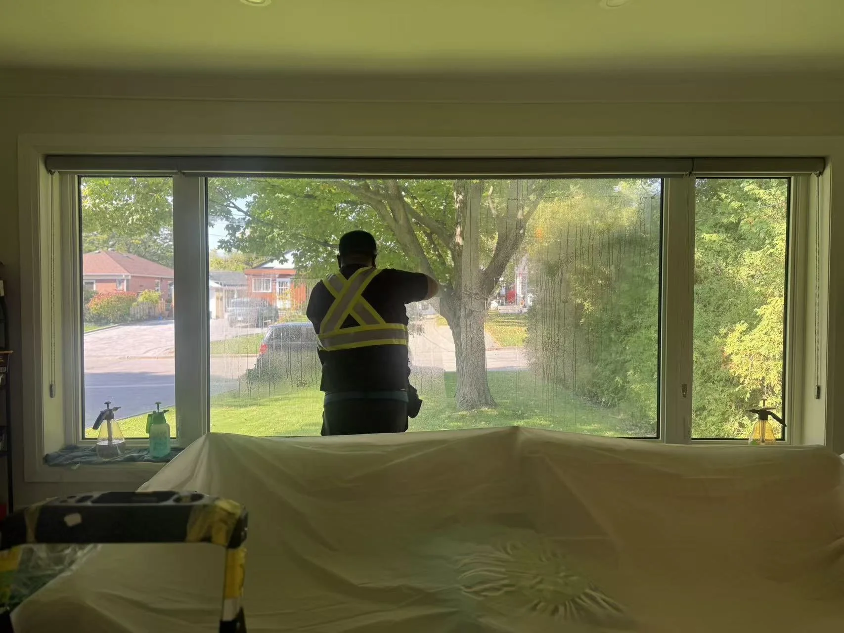 A person wearing a yellow safety vest cleaning or inspecting a large window from inside a house, with a view of a suburban neighborhood with trees and houses outside.