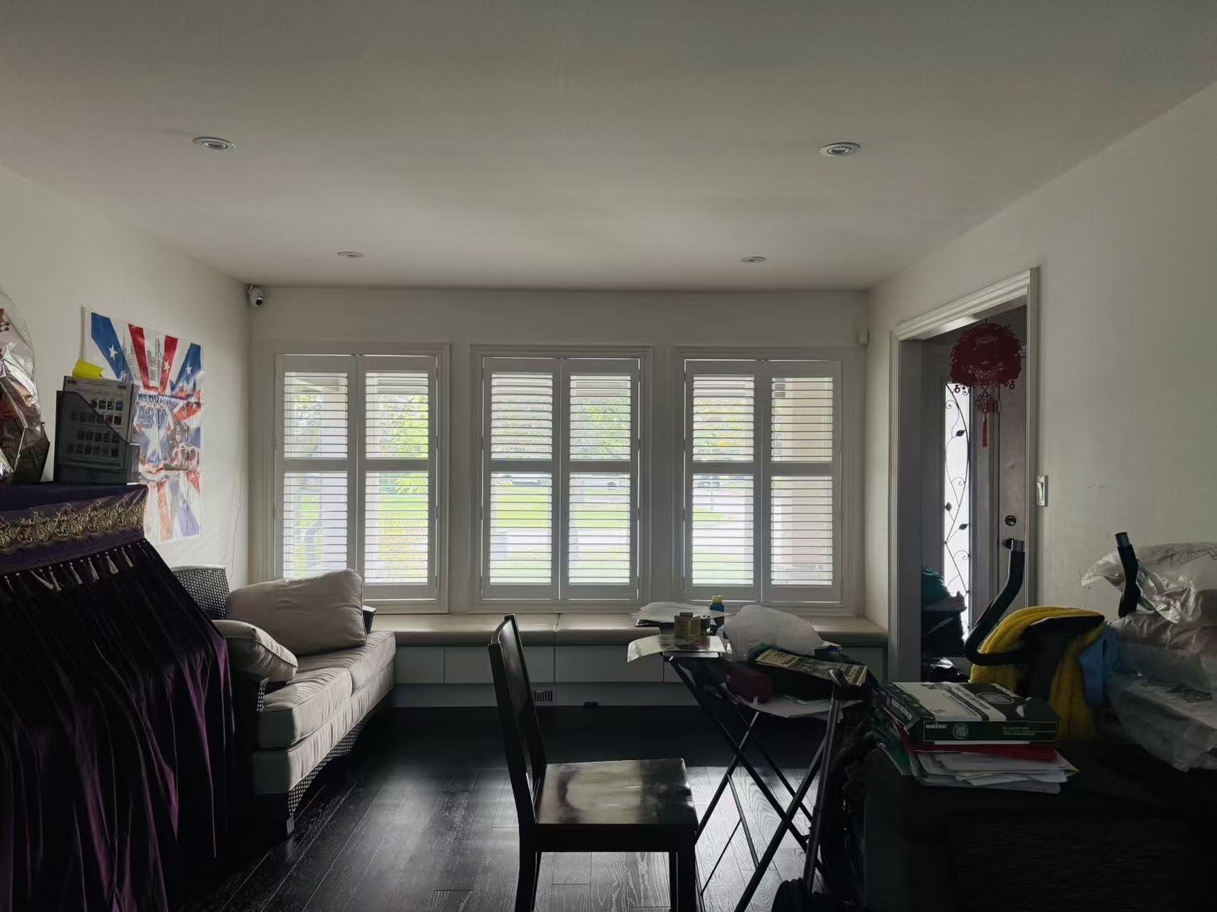 Room with three large windows with white shutters, a beige couch, a wooden chair, a cluttered table, and a door with a red decorative hanging.