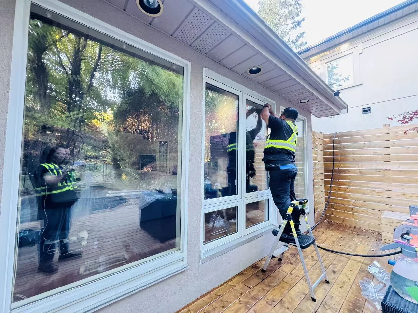 A person cleaning the exterior window of a house while standing on a ladder, with another person observing through the window. The scene is outdoor on a wooden deck, with a wooden fence on the right and trees reflected in the glass.