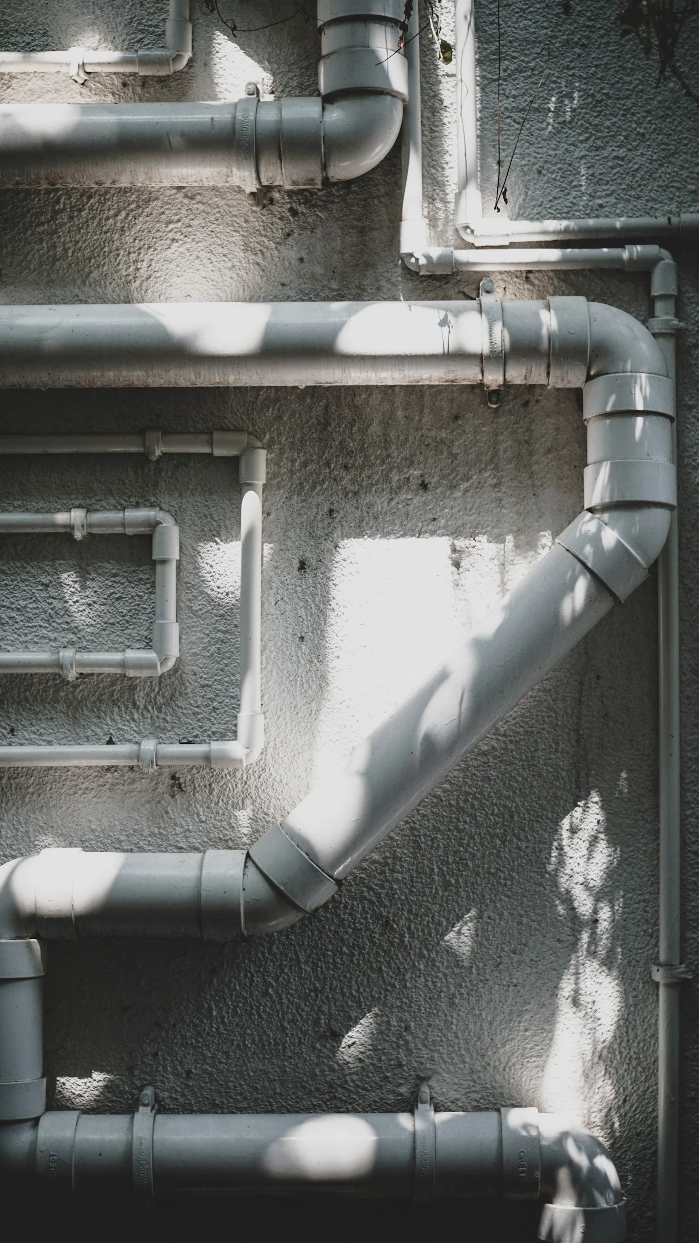Several white pipes of different sizes connected to a wall, with sunlight and shadows cast over them.