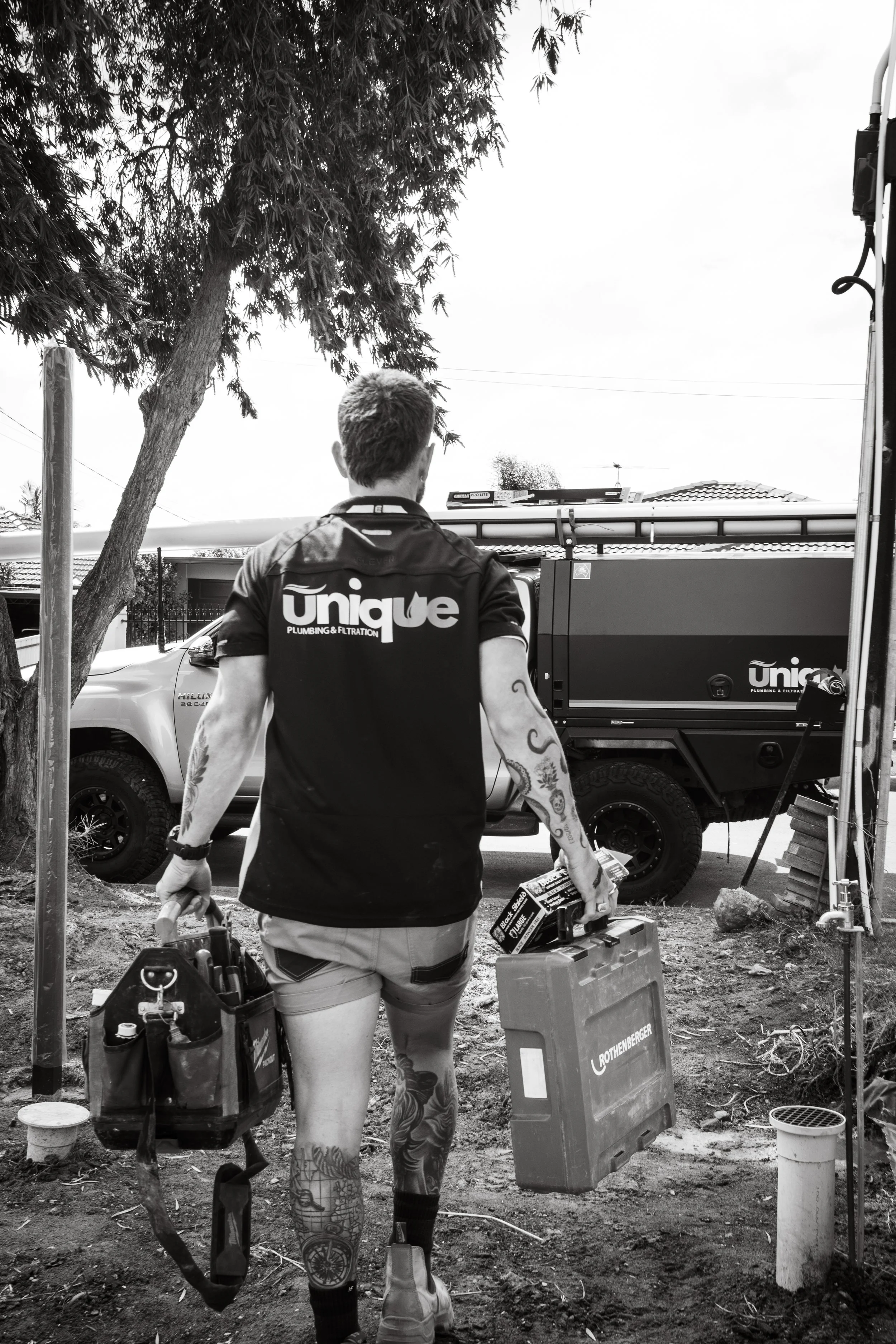 A man with tattoos on his legs and arms, wearing a black polo shirt with 'Unique Plumbing & Filtering' on the back, is carrying tools and a toolbox. He is walking towards a worksite with a truck and some equipment visible in the background.