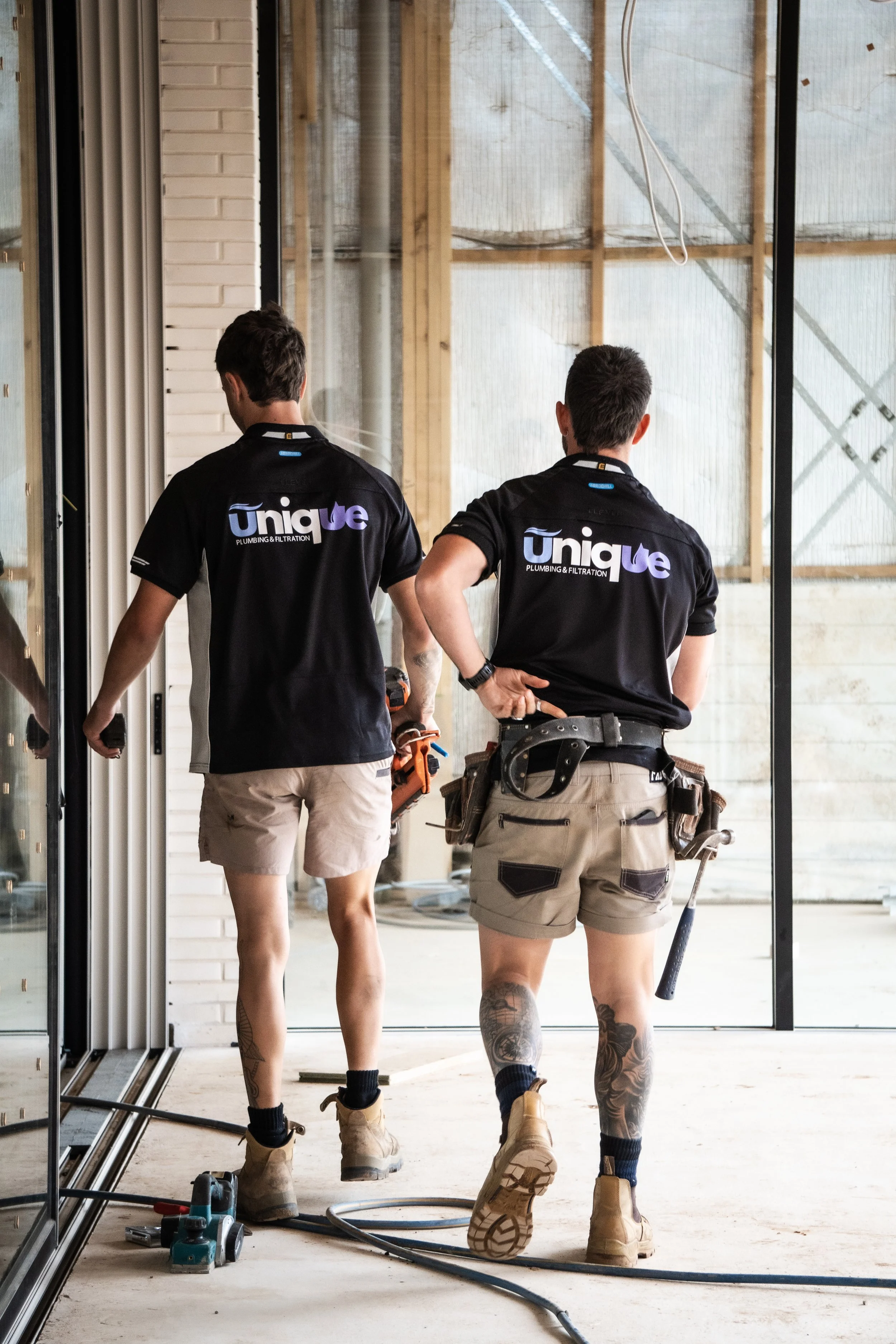Two construction workers in black shirts and tan shorts working inside a building under construction.
