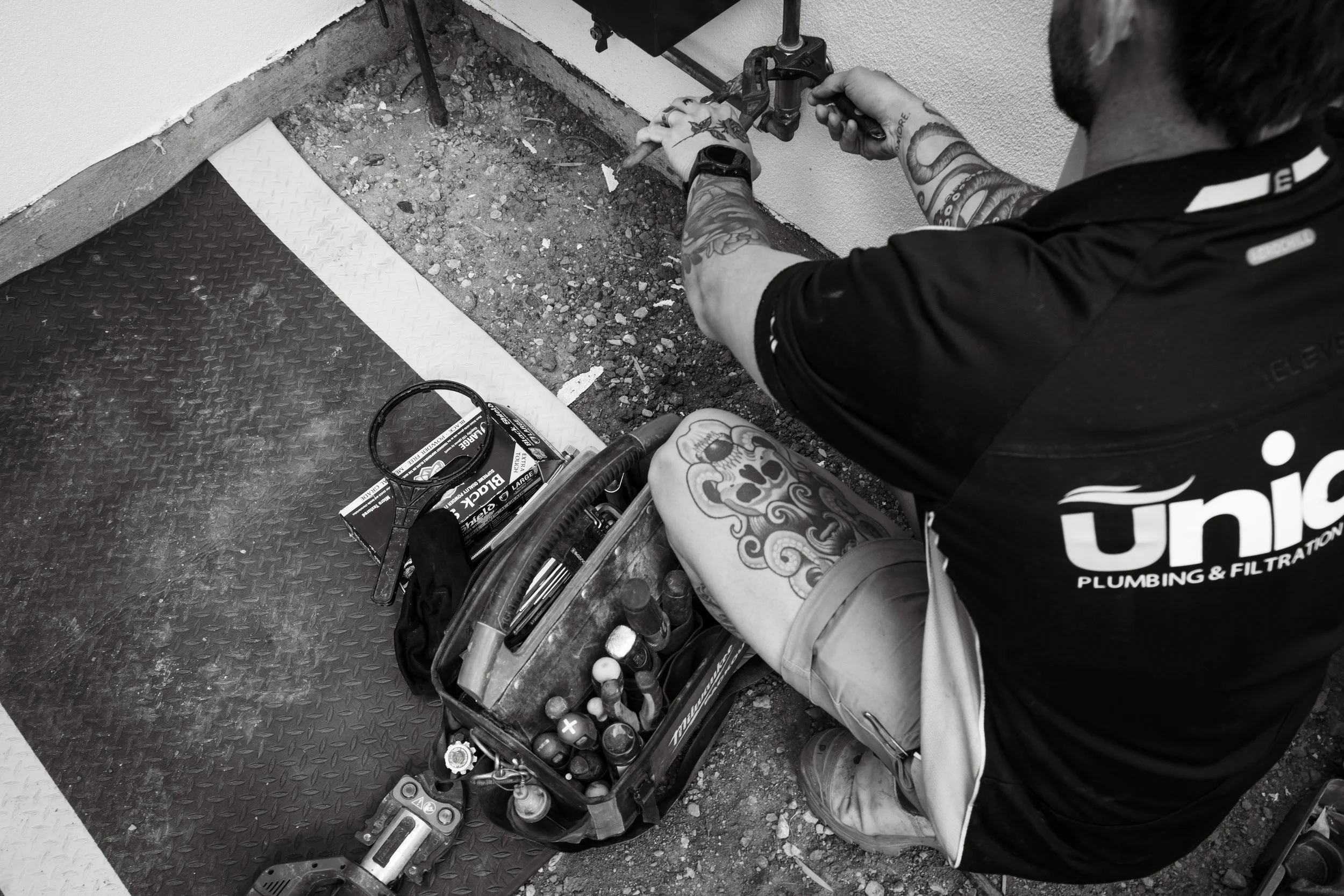 A plumbers working on pipes with tools and a toolbox on the ground, wearing a shirt with 'Union Plumbing & Filtration' logo.