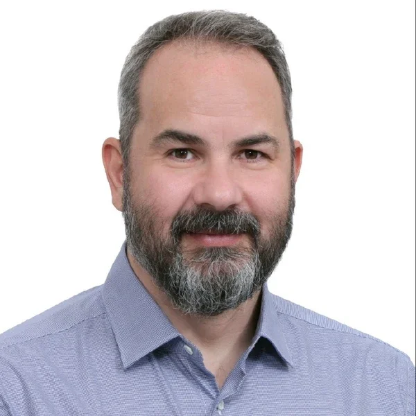 Headshot of a middle-aged man with a beard, smiling, wearing a light blue collared shirt against a plain white background.
