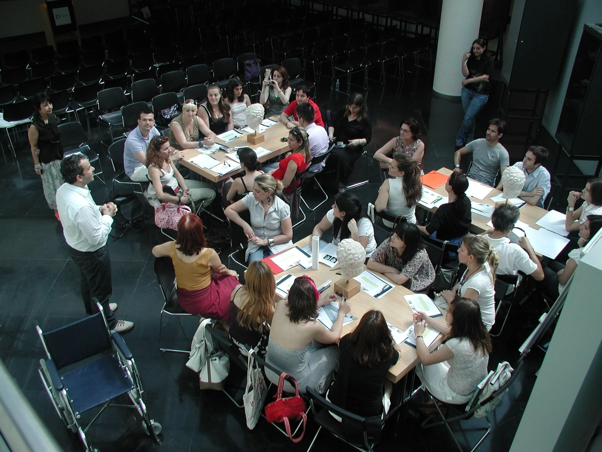 A group of people attending a meeting or discussion around a large rectangular table with bust sculptures and notebooks.