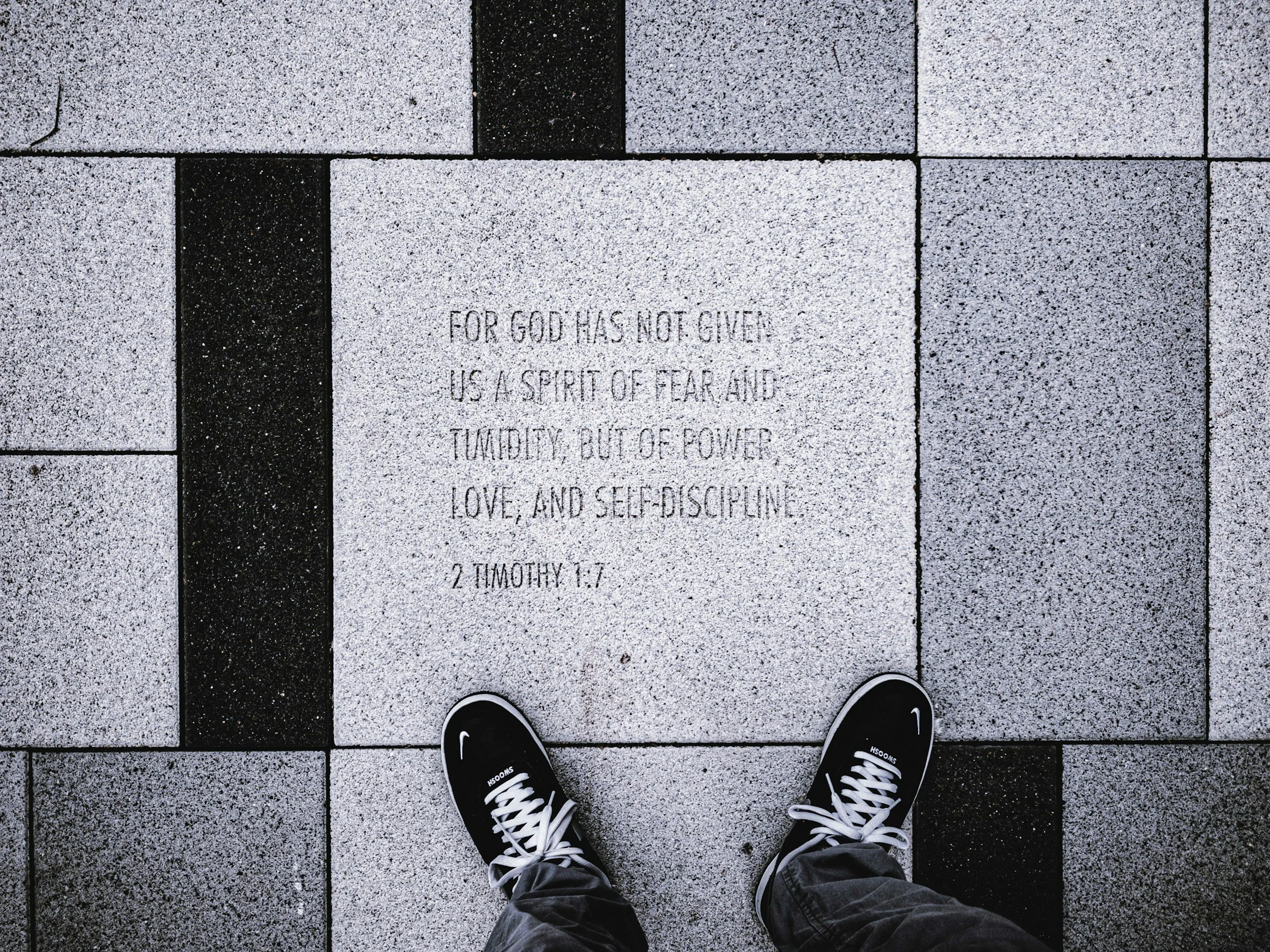 Person standing on a paved sidewalk, with the front of their feet and shoes visible, and an engraved quote on a stone tile reading, "For God has not given us a spirit of fear and humility, but of power, love, and self-discipline. 2 Timothy 1:7."