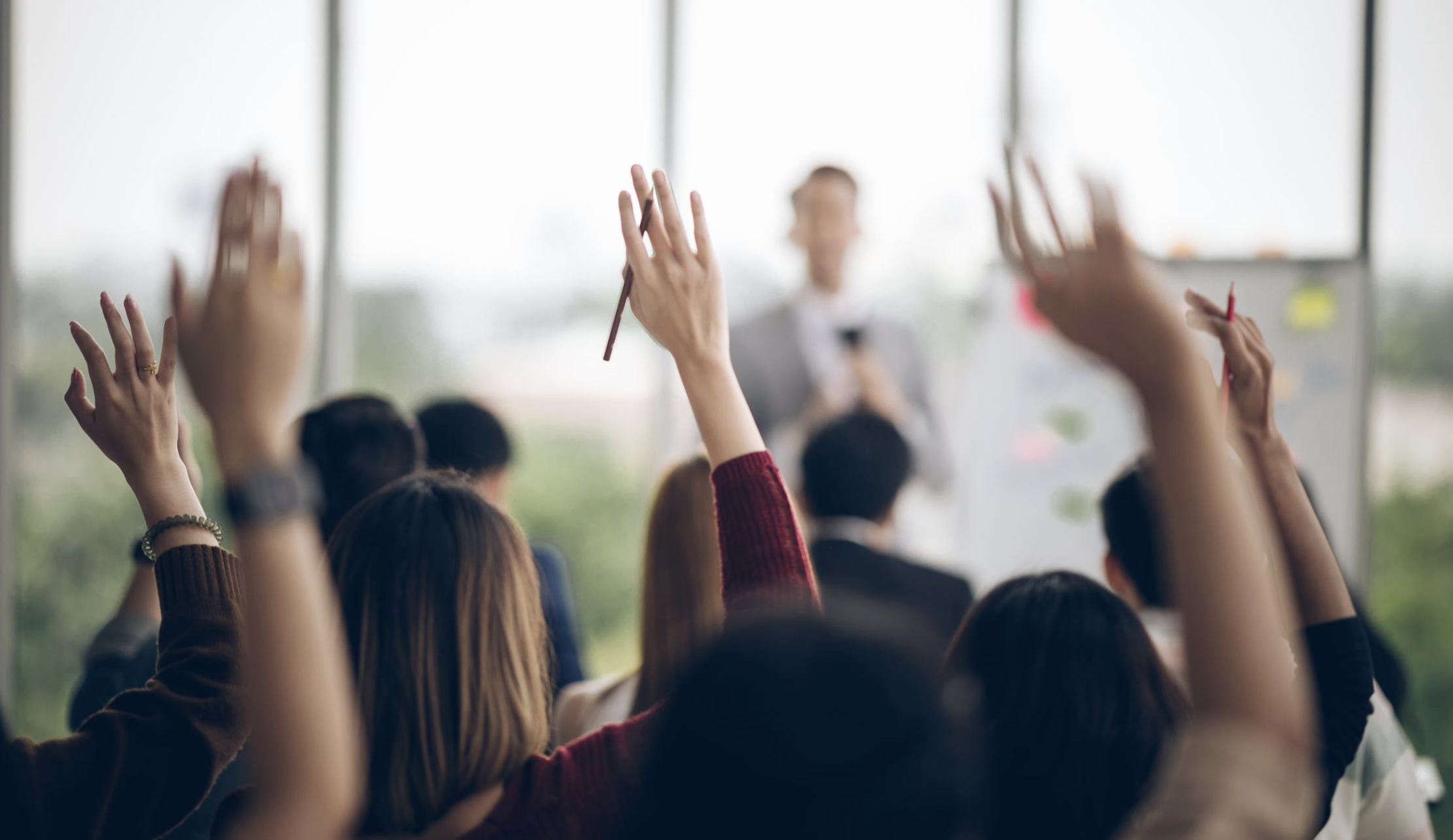 A classroom or conference room with many participants, some with hands raised, facing a speaker at the front with a whiteboard and large windows in the background