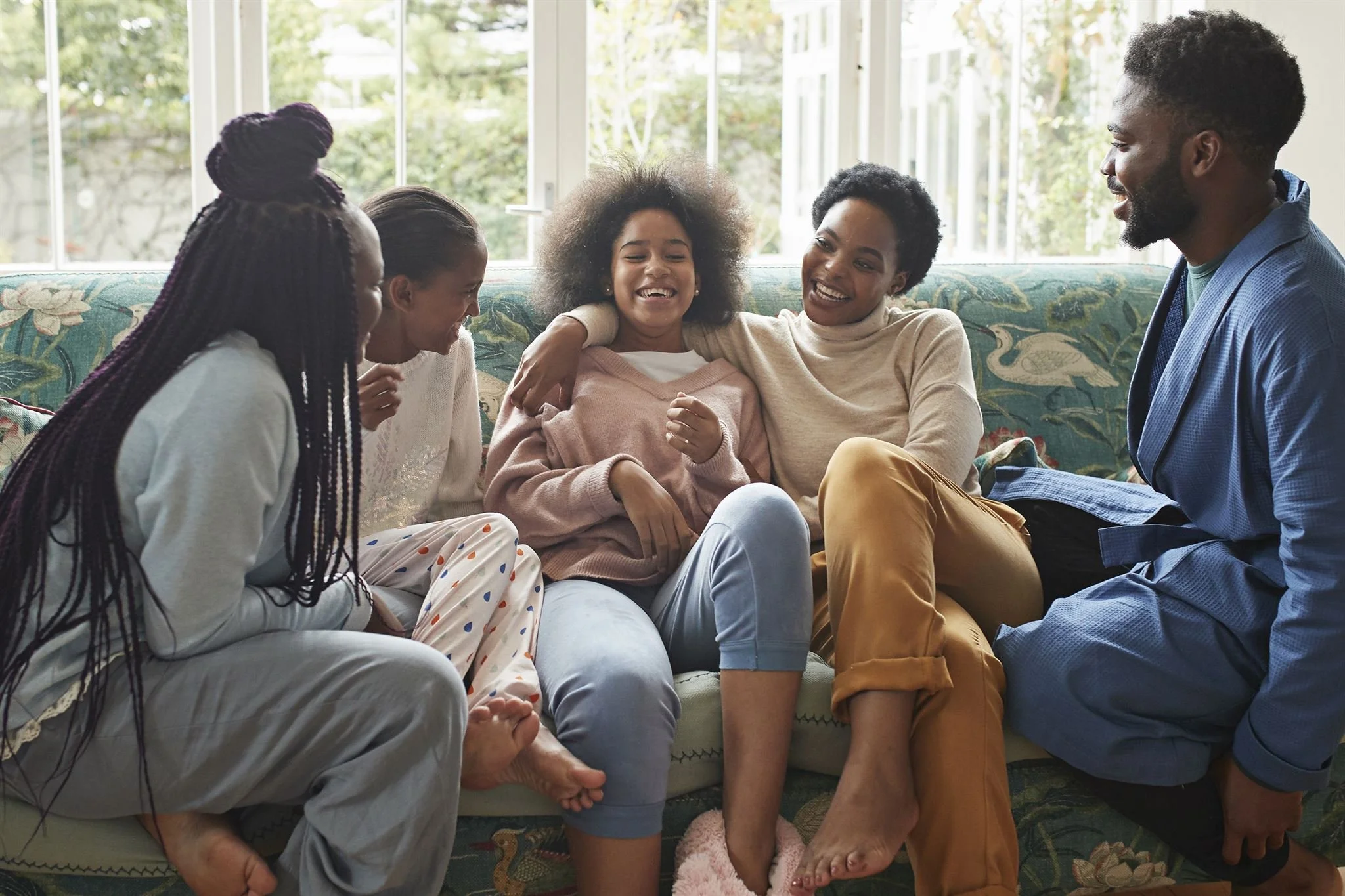 A diverse family of five, two adults and three children, sitting together on a sofa in a sunlit living room, sharing a joyful moment and laughing.
