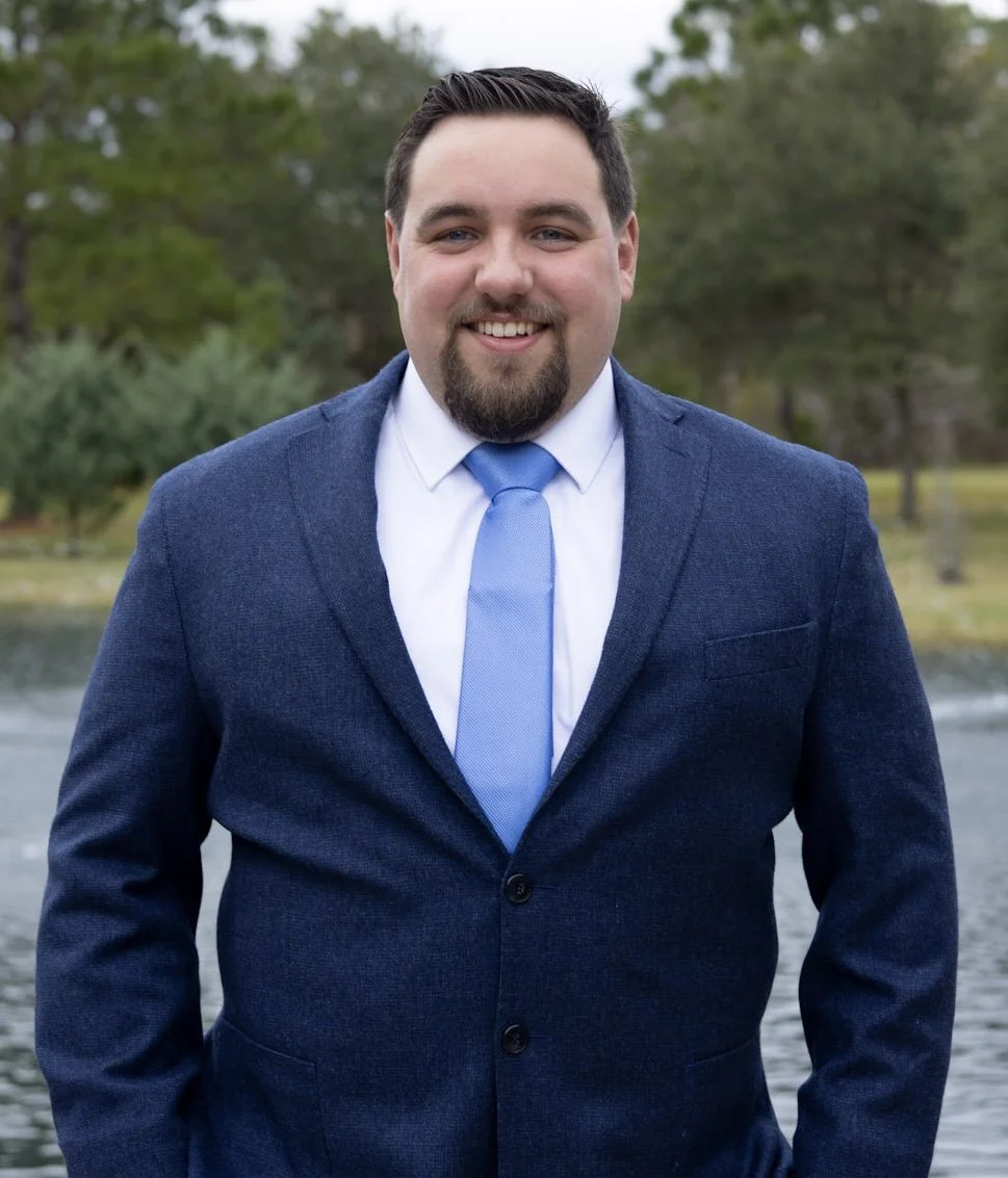 A man in a dark blue suit and light blue tie standing outdoors near a body of water with trees in the background.