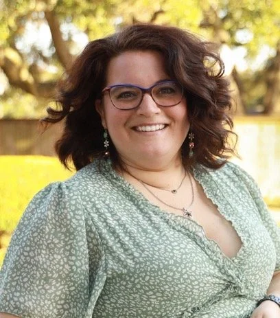A woman with curly brown hair, glasses, and earrings, smiling outdoors with trees and yellow flowers in the background.