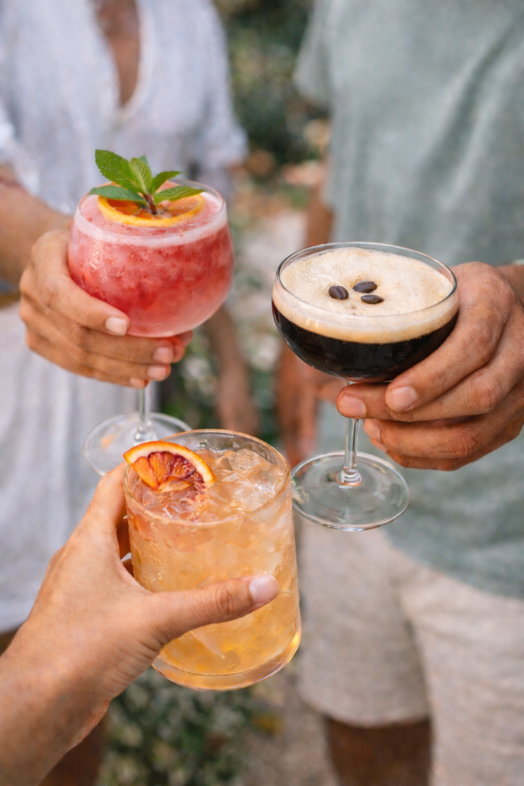 Three people holding different cocktails for a toast, with a pink drink garnished with mint leaves, a black coffee drink with coffee beans, and a peach-colored drink with blood orange slice.