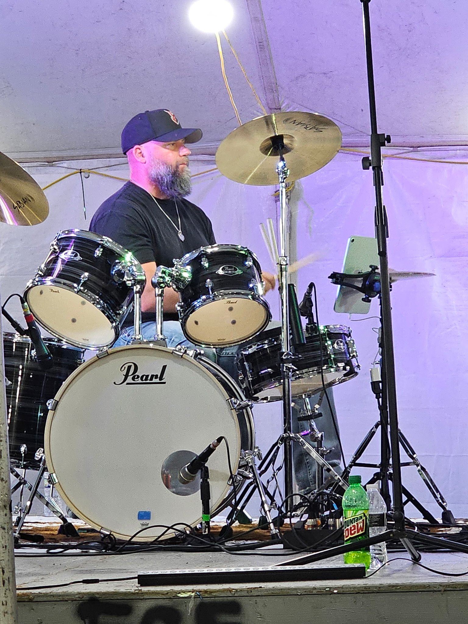 A man with a beard, wearing a black cap and black shirts, playing a drum set on stage with a white tent in the background. The drum set is black and white with the brand name Pearl, and there are drinks including Mountain Dew and a water bottle on the stage.
