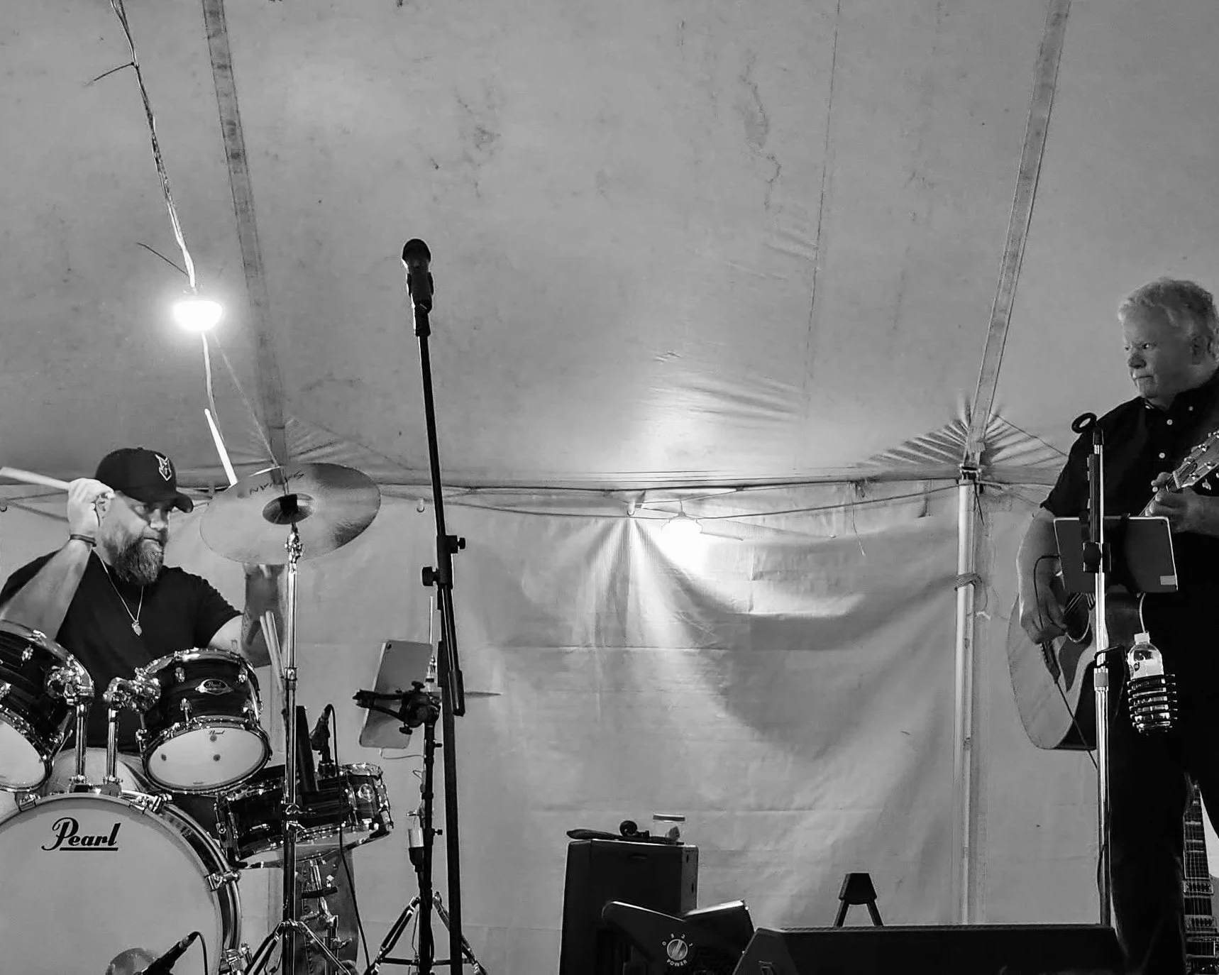 Two musicians performing on stage under a tent, one on drums and the other on guitar and vocals.