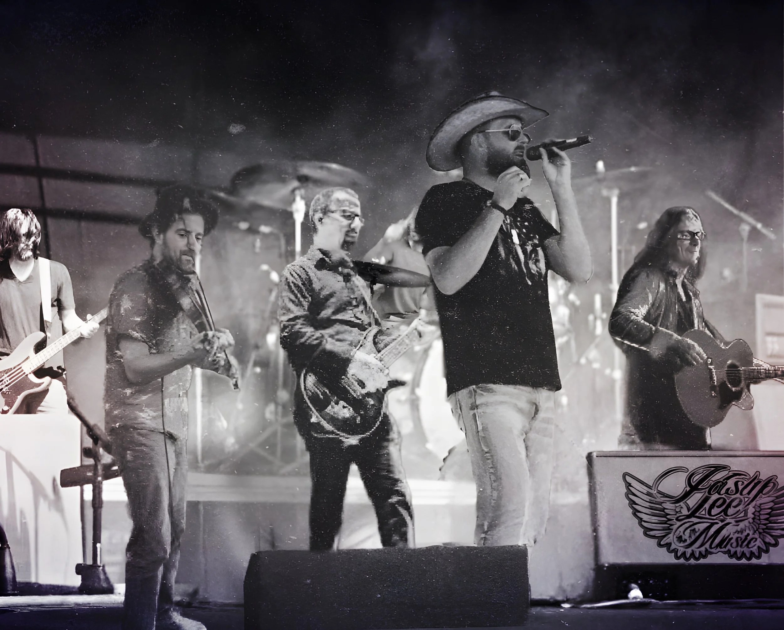 Black and white photo of a band performing on stage, with guitars, drums, and vocalists, including a man in a cowboy hat singing into a microphone.