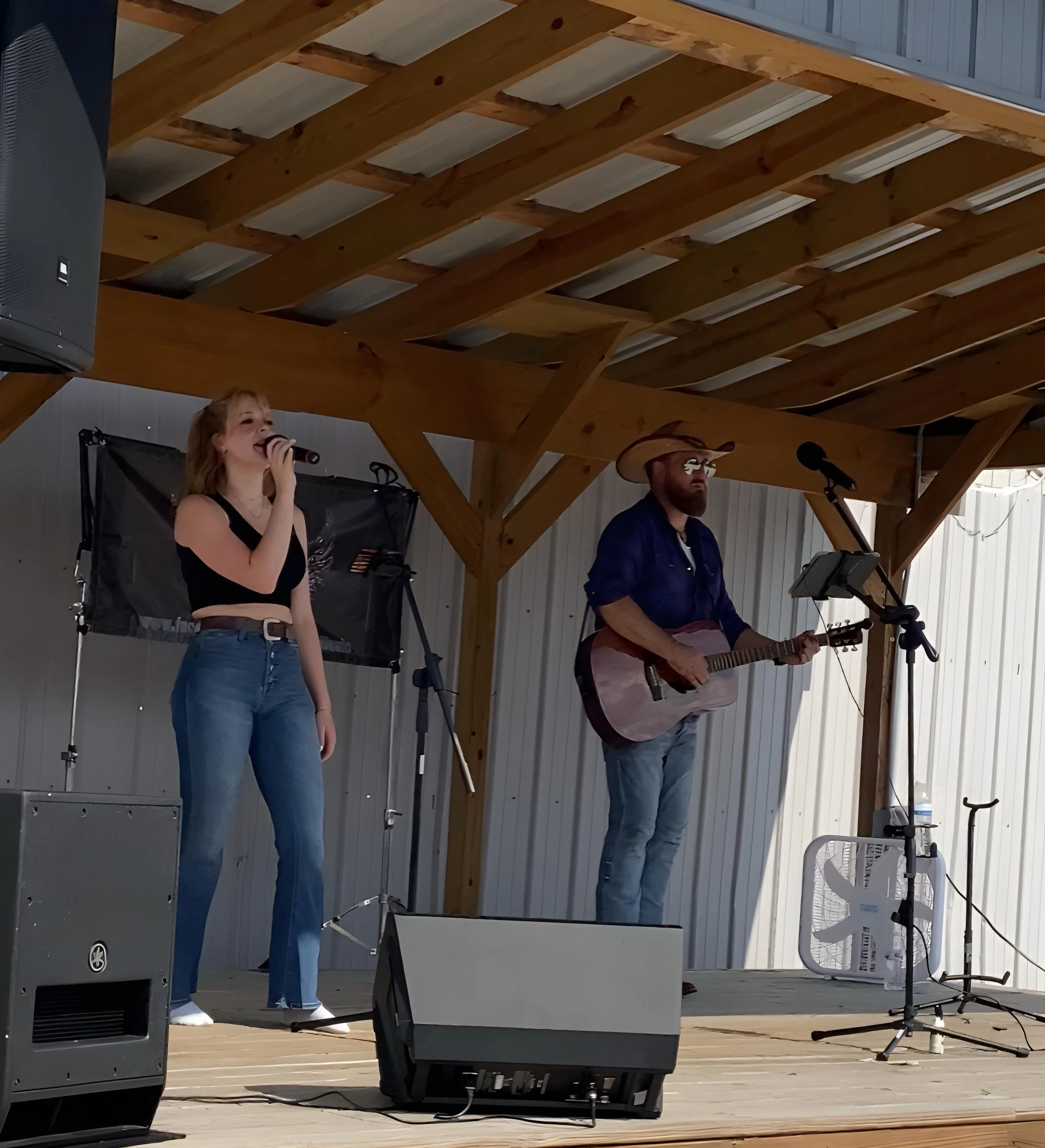 A young woman singing into a microphone on a wooden stage, accompanied by a man playing guitar, under a wooden roof structure.