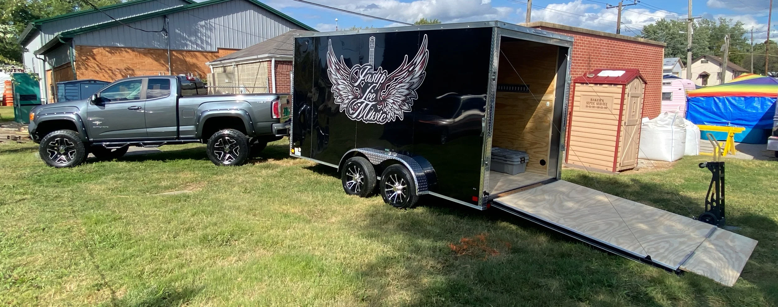Gray pickup truck parked next to a black trailer with angel wings and a guitar logo, on a grassy lot with buildings and tents in the background.