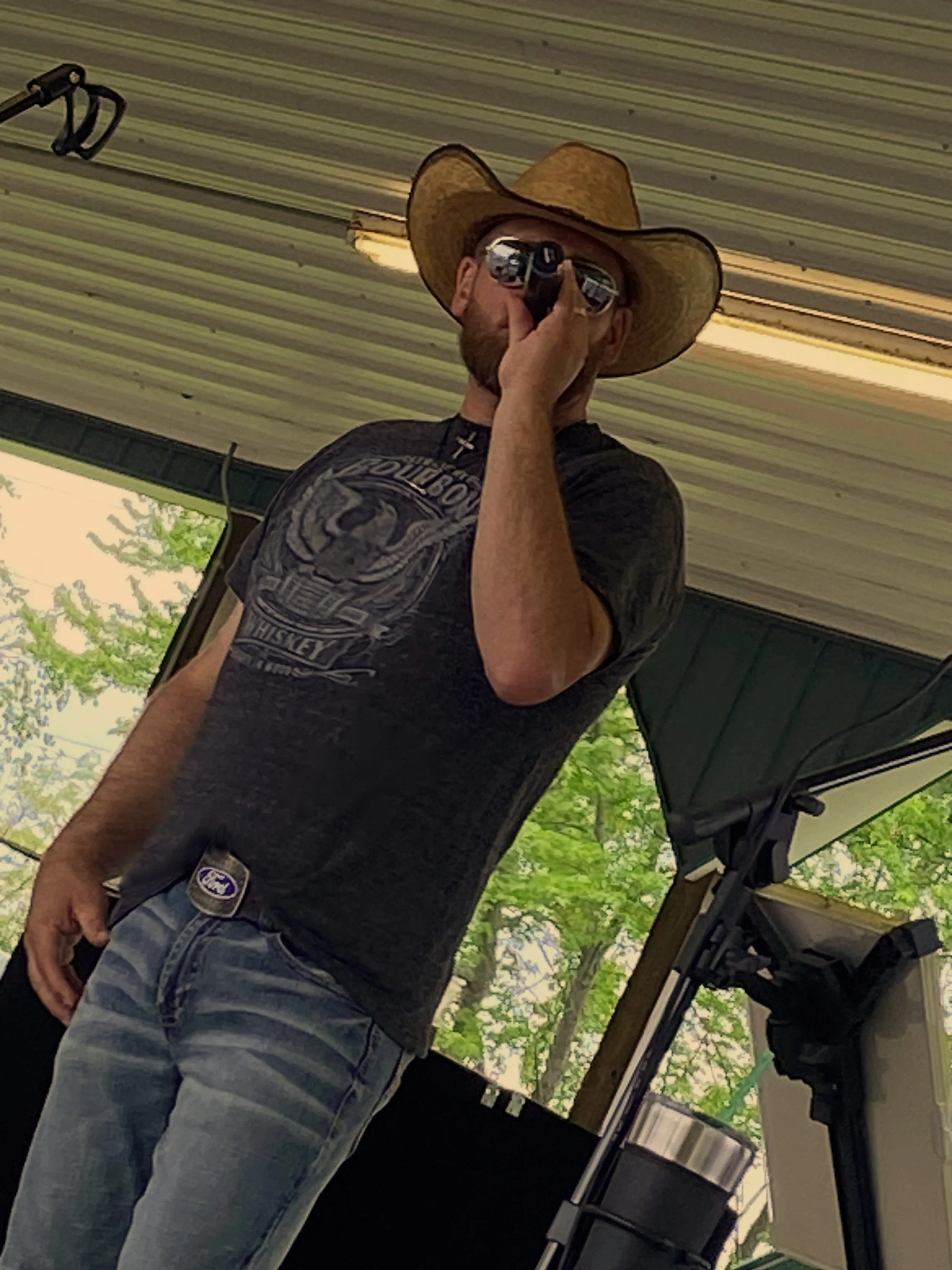 A man wearing a cowboy hat, sunglasses, a black graphic t-shirt, and jeans, standing under a metal roof, taking a selfie with his phone.