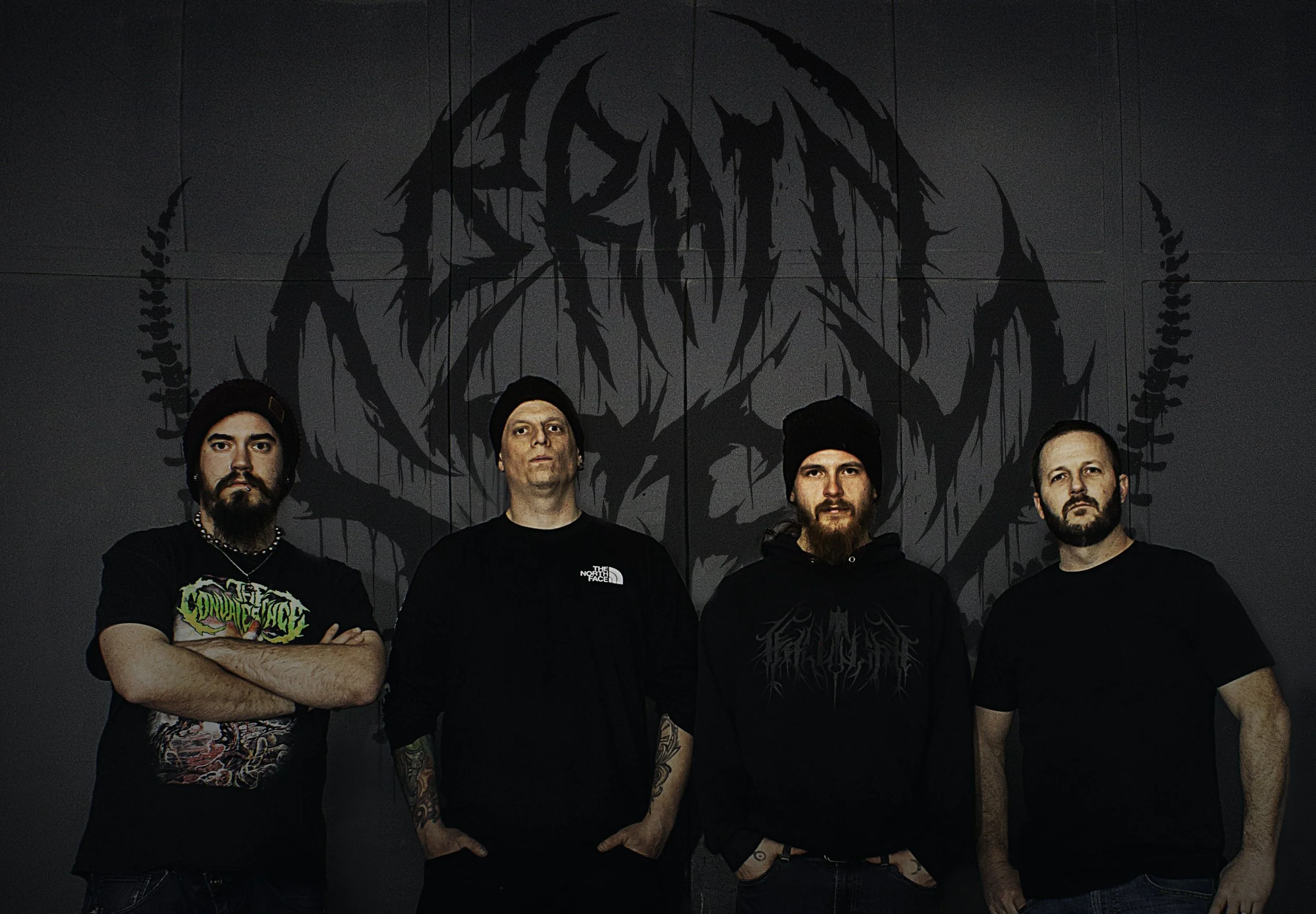 Four men standing in front of a black wall with a large dark logo. The men are wearing black hoodies, t-shirts, and beanies, with one having tattoos visible on his arms. They have serious expressions and are looking at the camera.