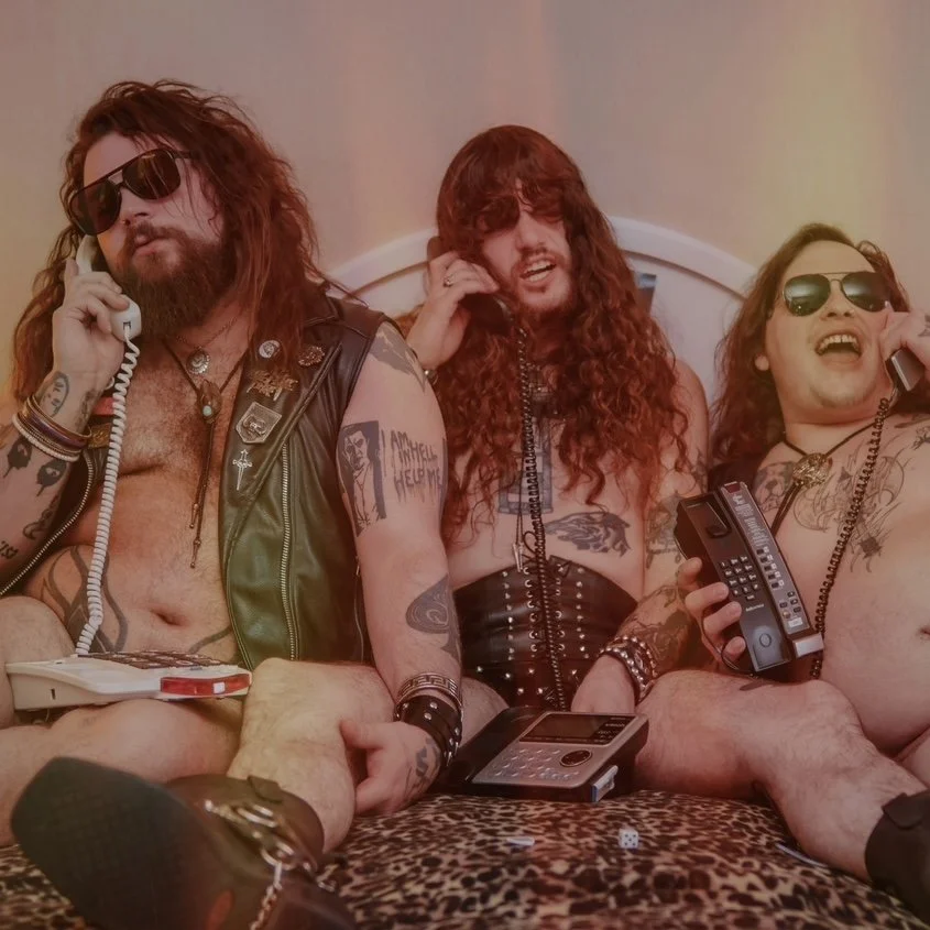 Three men with long hair and sunglasses sitting on a bed, holding phones, with various tattoos and punk attire.