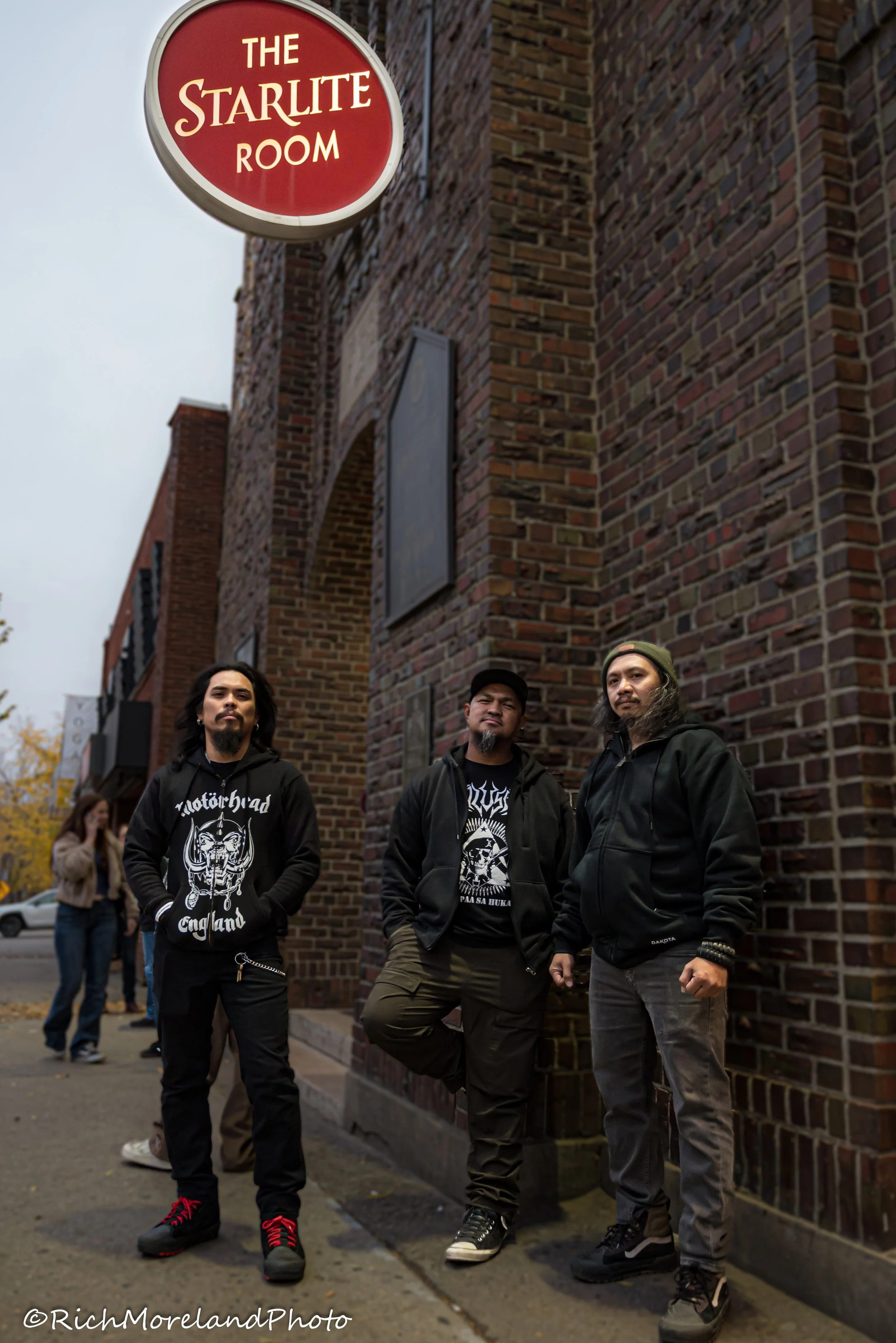 Three men standing outside the brick entrance of The Starlite Room, a music venue, on a cloudy day, with a woman and a car in the background.