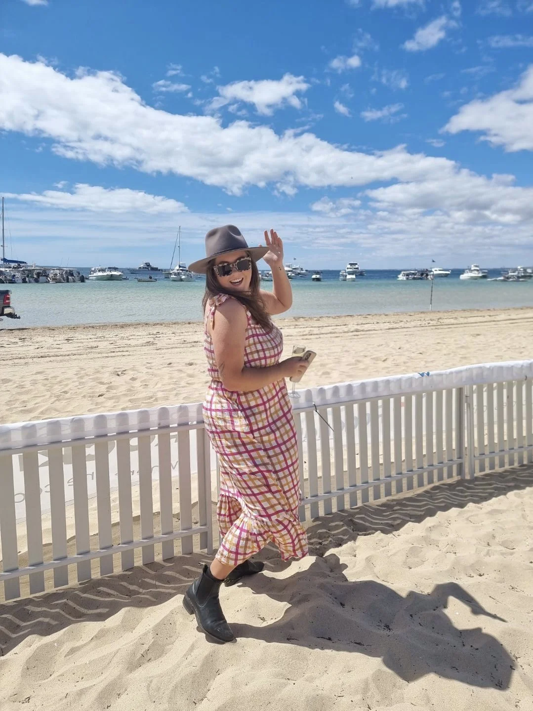 A woman in a plaid dress, wide-brimmed hat, sunglasses, and black boots standing on a sandy beach by a white fence, waving and smiling at the camera. The background features boats docked on the water under a blue sky with scattered clouds.