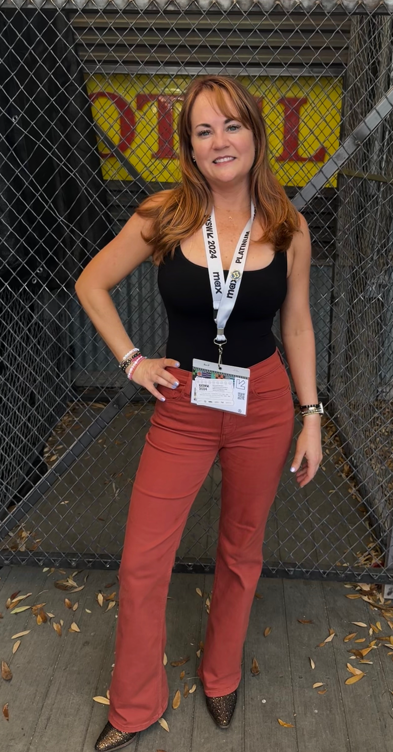 A woman with red hair standing in front of a chain-link fence, wearing a black tank top, red pants, and glittery shoes, with event badges around her neck.