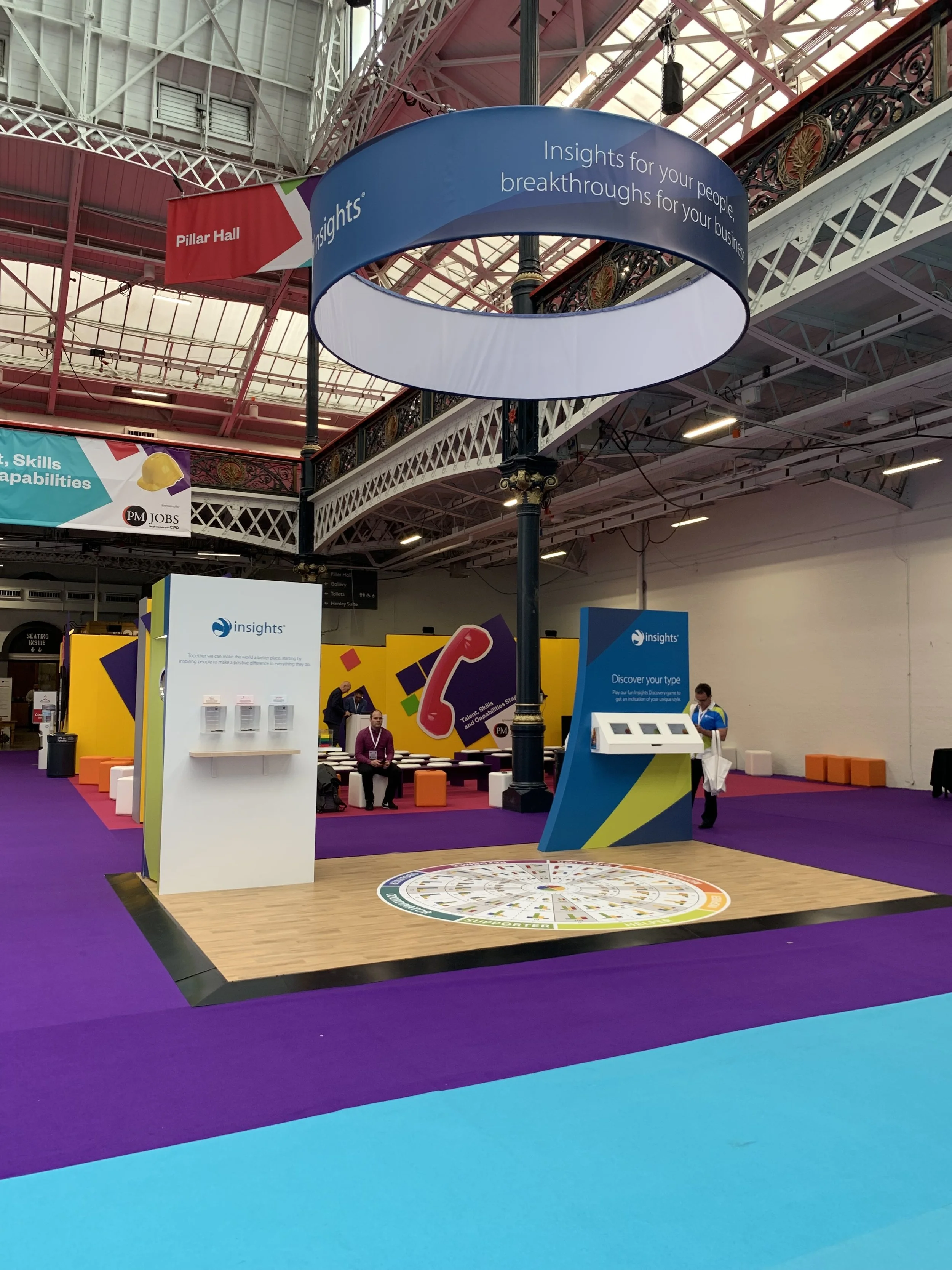A convention hall booth for Insights features colorful signage, a circular display with a wheel, and informational panels, with purple, blue, and yellow accents. There are a few visitors and staff members in the area.